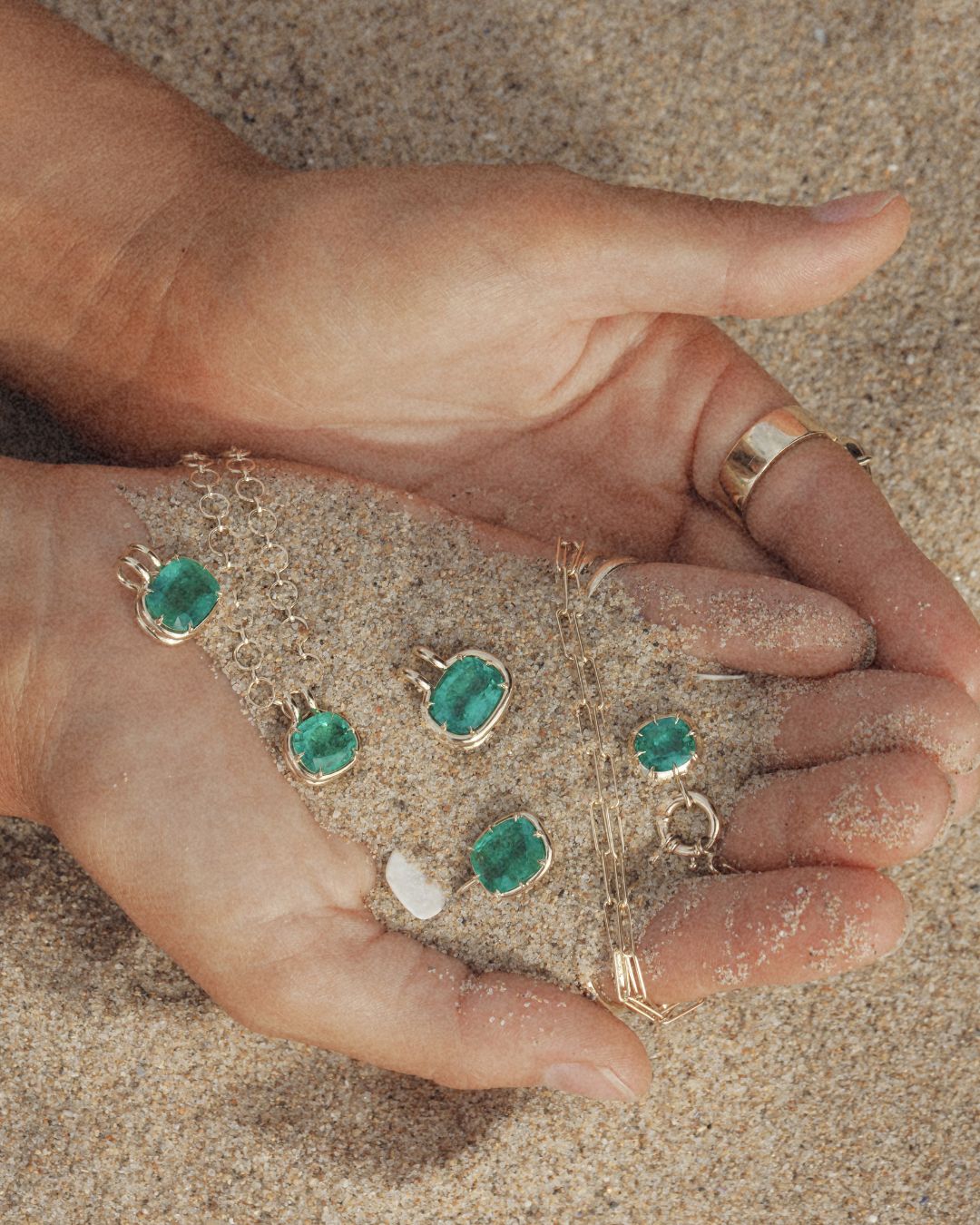 Gold jewelry pieces, including a necklace and rings, displayed on sand held in two hands.