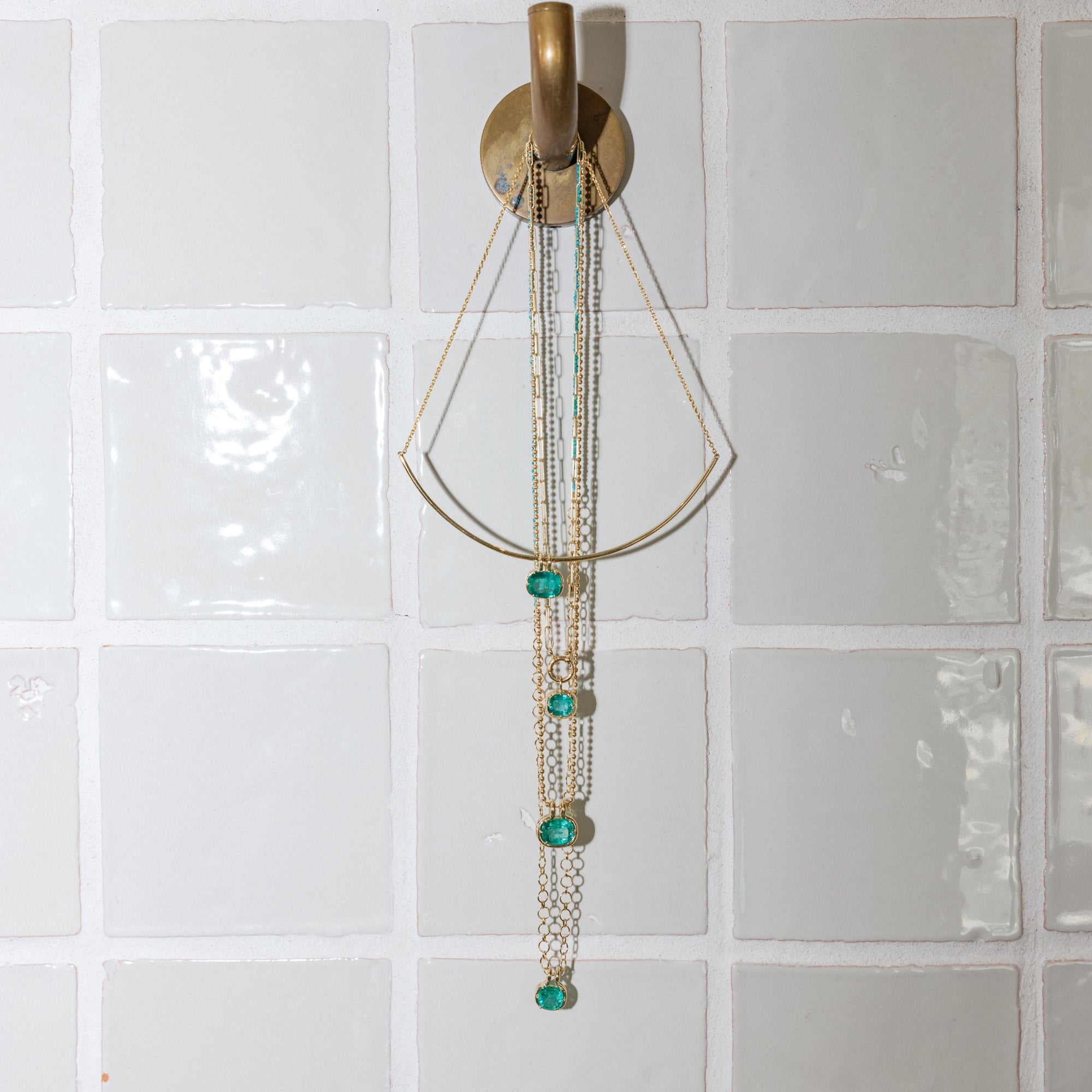 A gold-toned wall-mounted jewelry organizer with multiple hanging chains and several emerald green gemstone accents that would match perfectly with unique engagement rings by Sofia Kaman. 