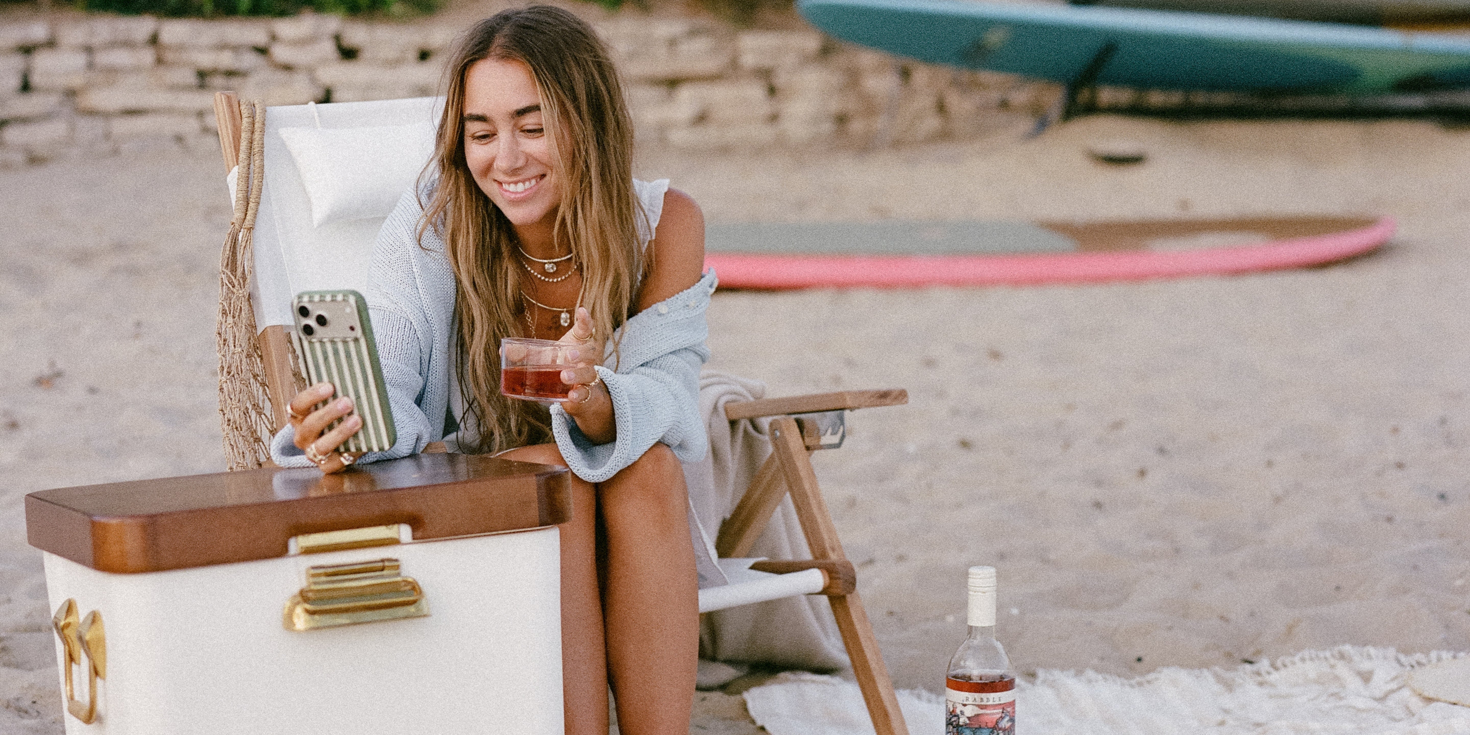 A woman sitting in a beach chair on a sandy beach, holding a drink and looking at a phone. A white and wood cooler with gold hardware is in the foreground, with a bottle on a blanket nearby and surfboards in the background.