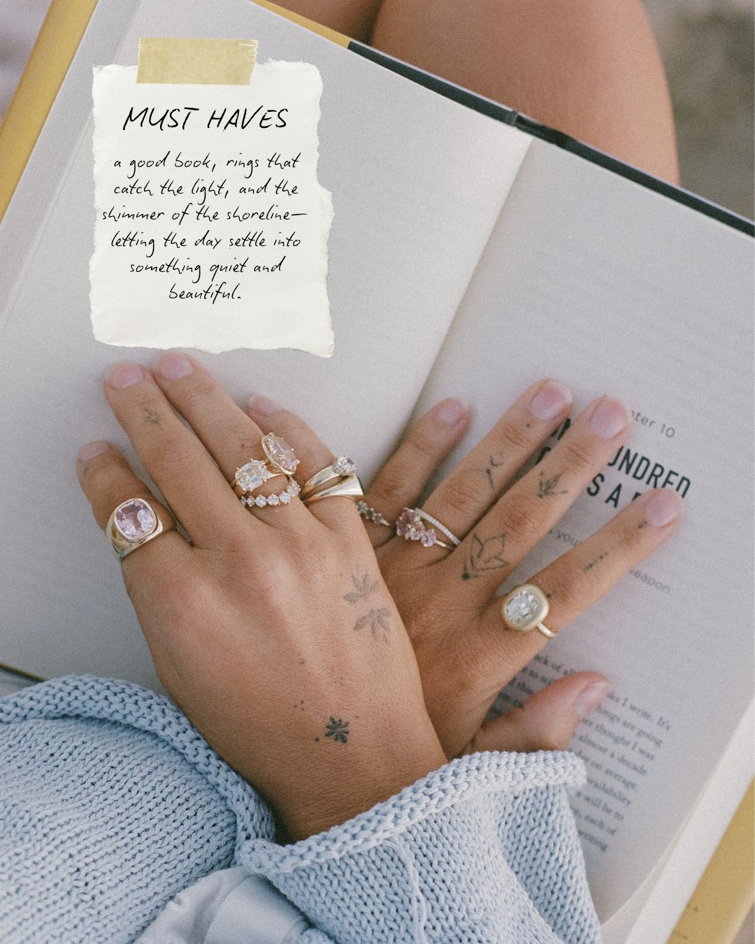 A close-up of a person's hands resting on an open book, wearing multiple delicate rings with diamonds and soft pink accents. A handwritten note on the book reads 'MUST HAVES' and lists ring details.