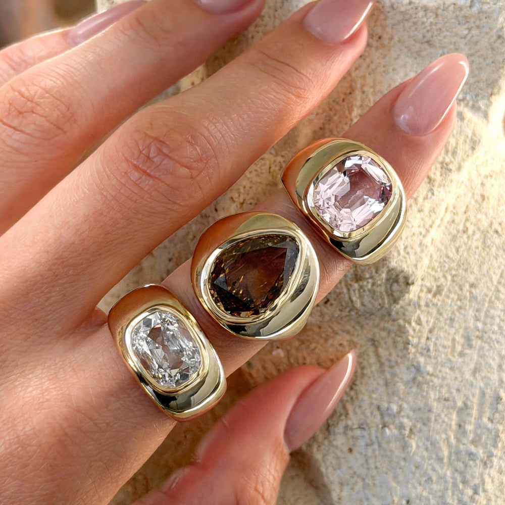 Hand wearing three gold rings with different colored gemstones on a neutral background