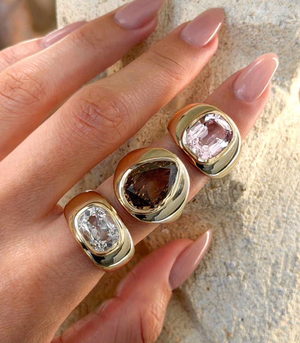 Hand wearing three gold rings with different colored gemstones on a neutral background