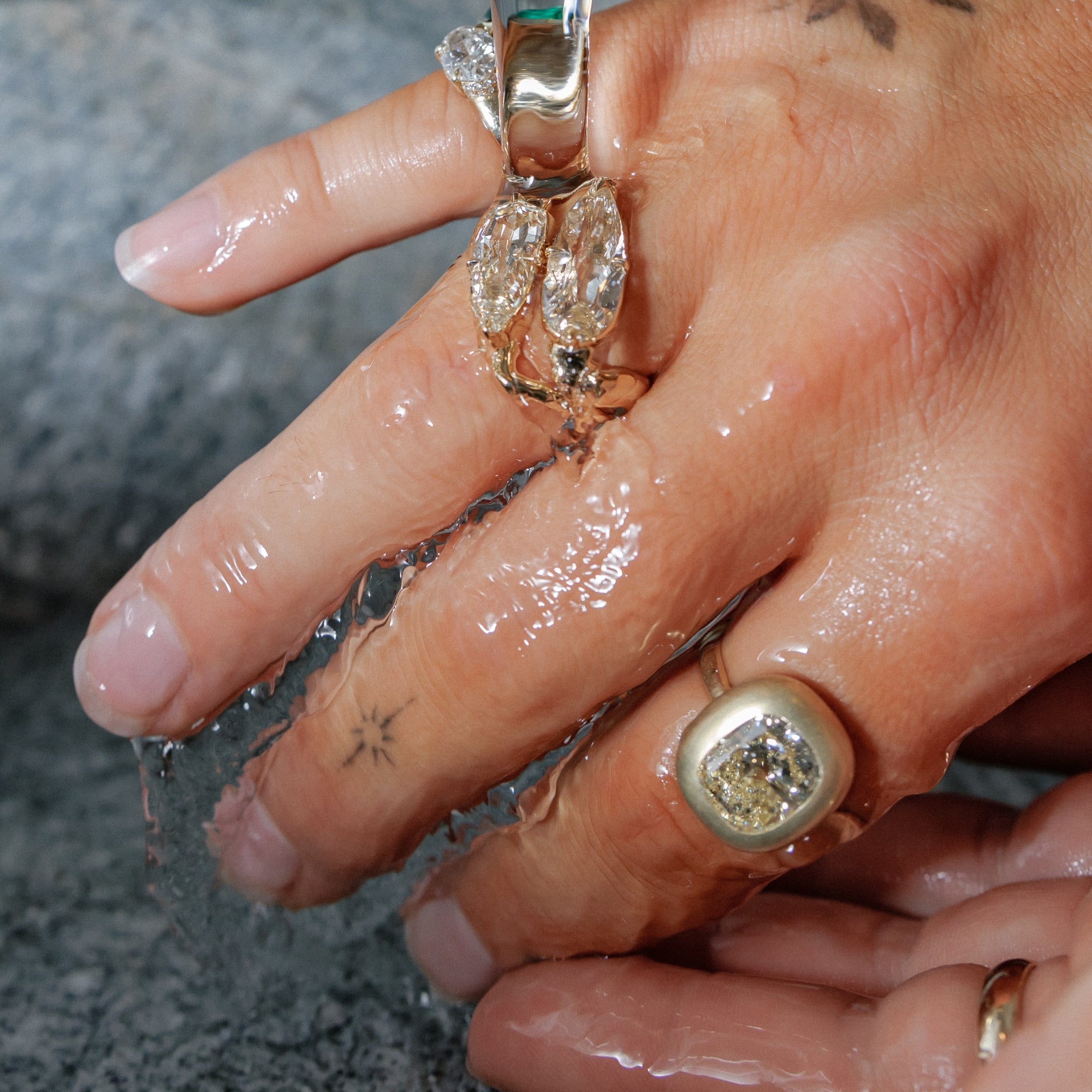 A close-up of a hand under running water, wearing multiple rings including a prominent gold ring with a large clear diamond and a gold ring with two clear marquise-cut diamonds.