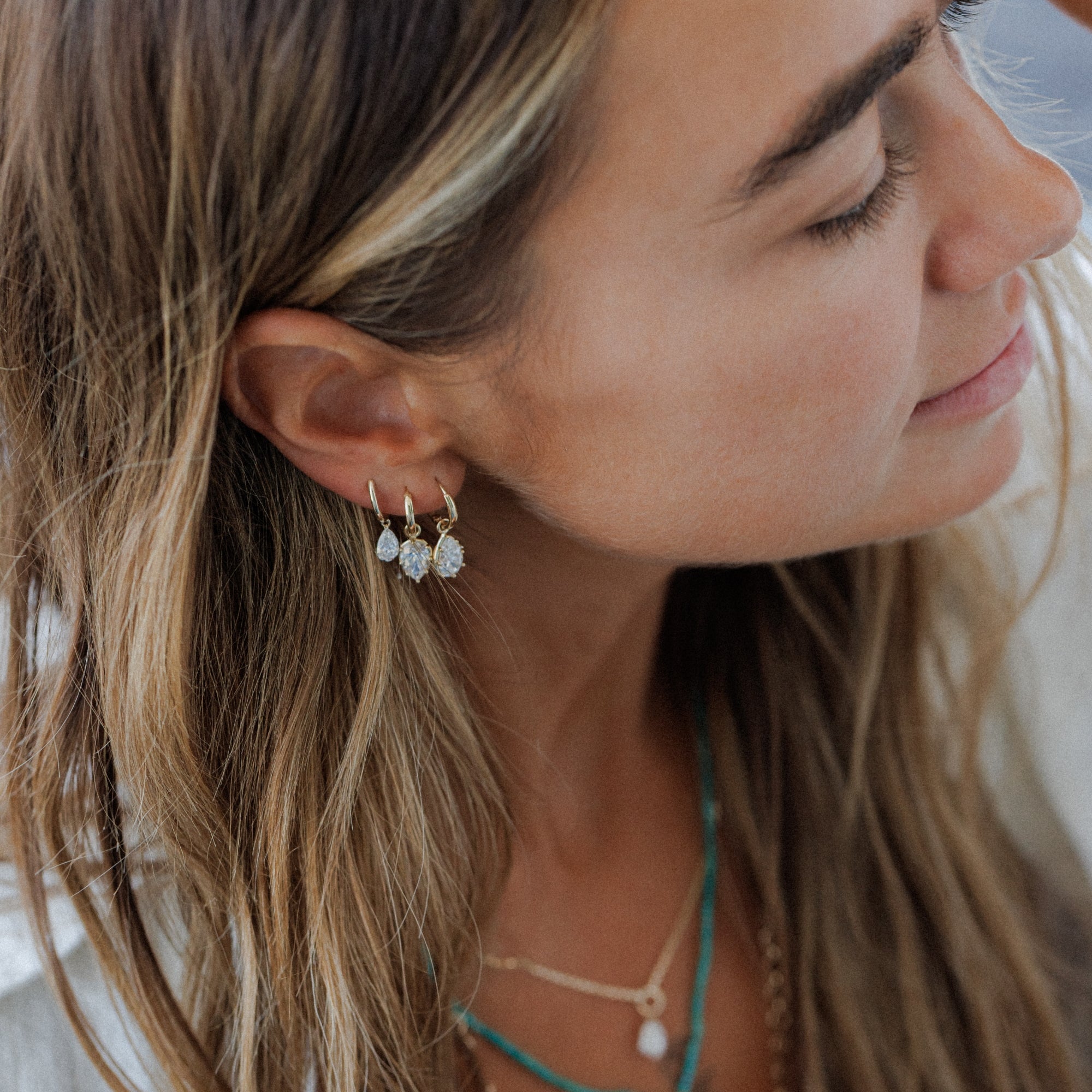 A close-up side profile of a woman with long light brown hair wearing small gold dangle earrings with white bead details, layered with turquoise and gold necklaces.