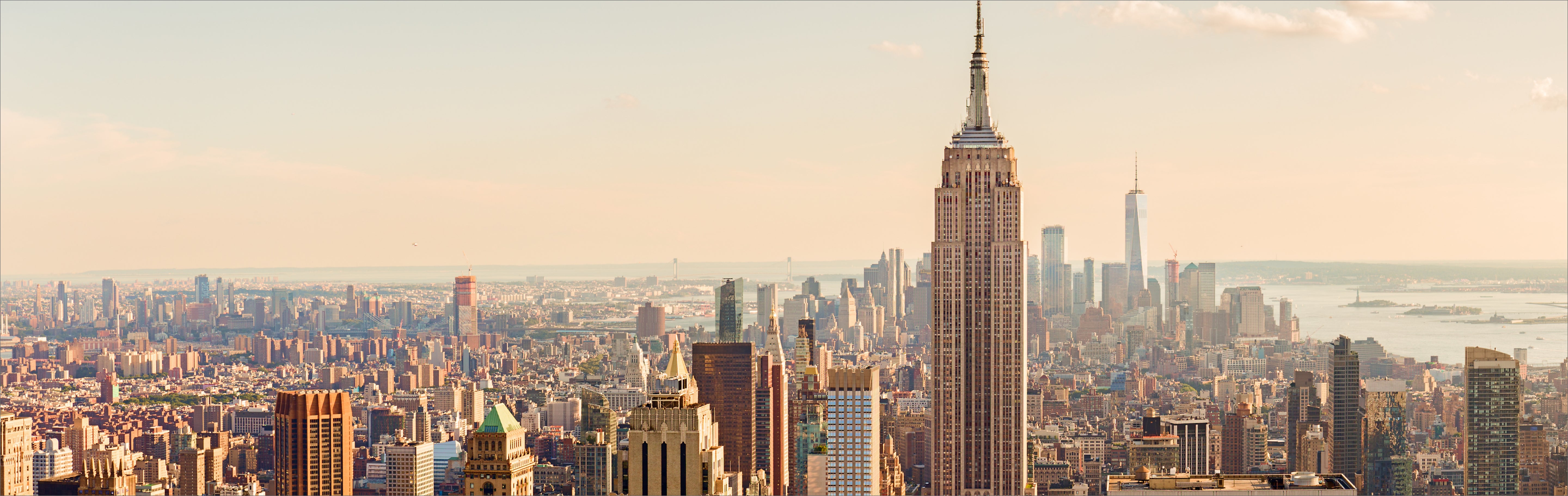Wide city skyline at sunset with tall skyscrapers, the Empire State Building centerpiece, and distant harbor.