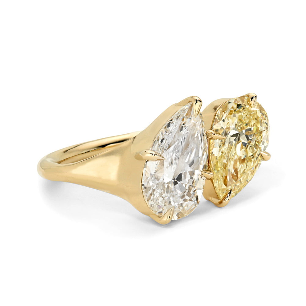 A yellow gold ring featuring two pear brilliant-cut diamonds, one white and one yellow, set in a polished gold band against a white background.
