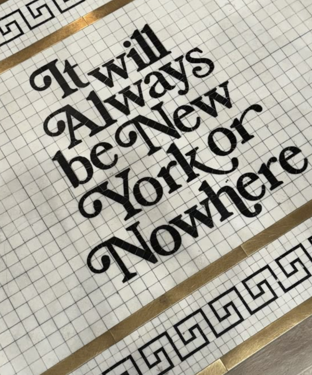 A square decorative tile or floor mat with the phrase 'It will always be New York or Nowhere' in black script, bordered by black Greek key motifs and metallic gold stripes on a light beige grid background.