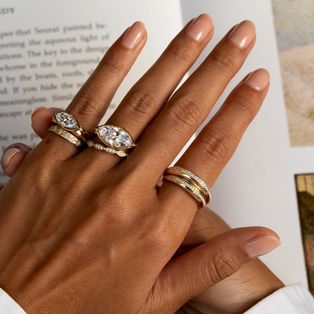 Close-up of a hand with a book in the background, styled with yellow gold and diamond rings, featuring two east-west marquise diamond rings paired with delicate diamond bands and a layered pavé pinky ring stack.