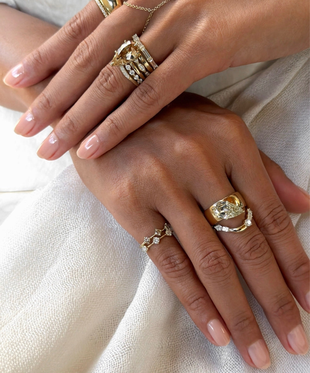 Close-up of two hands styled with layered yellow gold and diamond rings, featuring pear-shaped champagne and white diamond stacking bands, sculptural gold pear shaped band, delicate diamond pinky rings, and a fine gold hand chain.