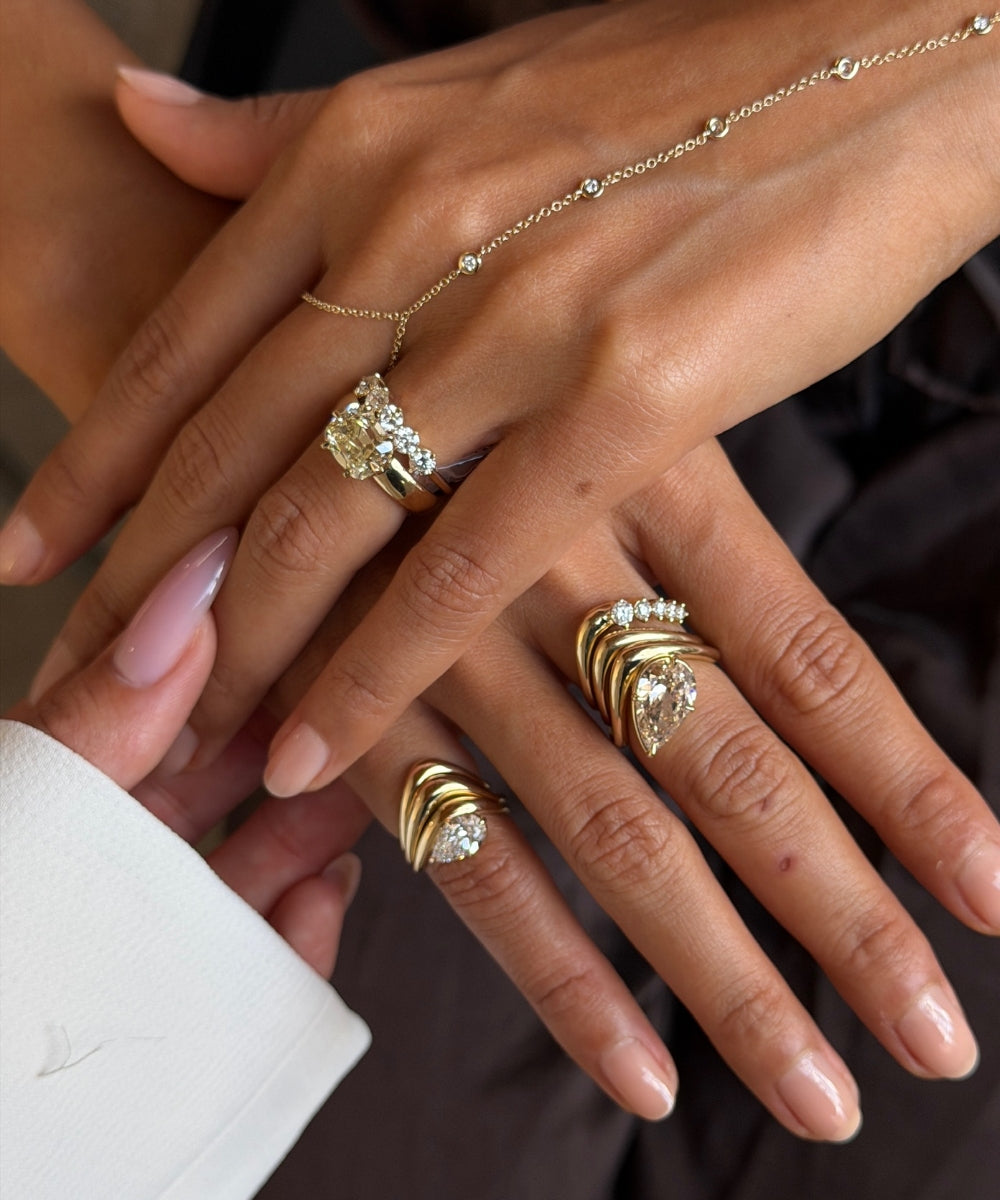 A close-up of two hands displaying a collection of gold rings and a delicate gold chain necklace. The jewelry features various designs including a large cluster of clear stones, a twisted band ring, and a smaller twisted band ring, set against a dark background.
