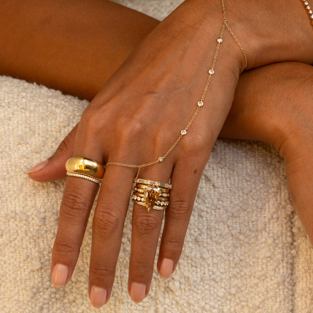 Close-up of a hand styled with layered yellow gold and diamond jewelry, featuring a marquise champagne diamond ring stack, a sculptural gold pinky ring, delicate diamond bands, and a fine diamond hand chain.