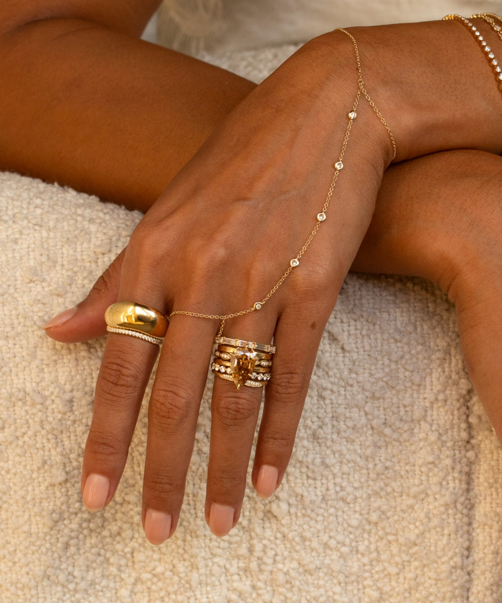 Close-up of a hand styled with layered yellow gold and diamond jewelry, featuring a marquise champagne diamond ring stack, a sculptural gold pinky ring, delicate diamond bands, and a fine diamond hand chain.