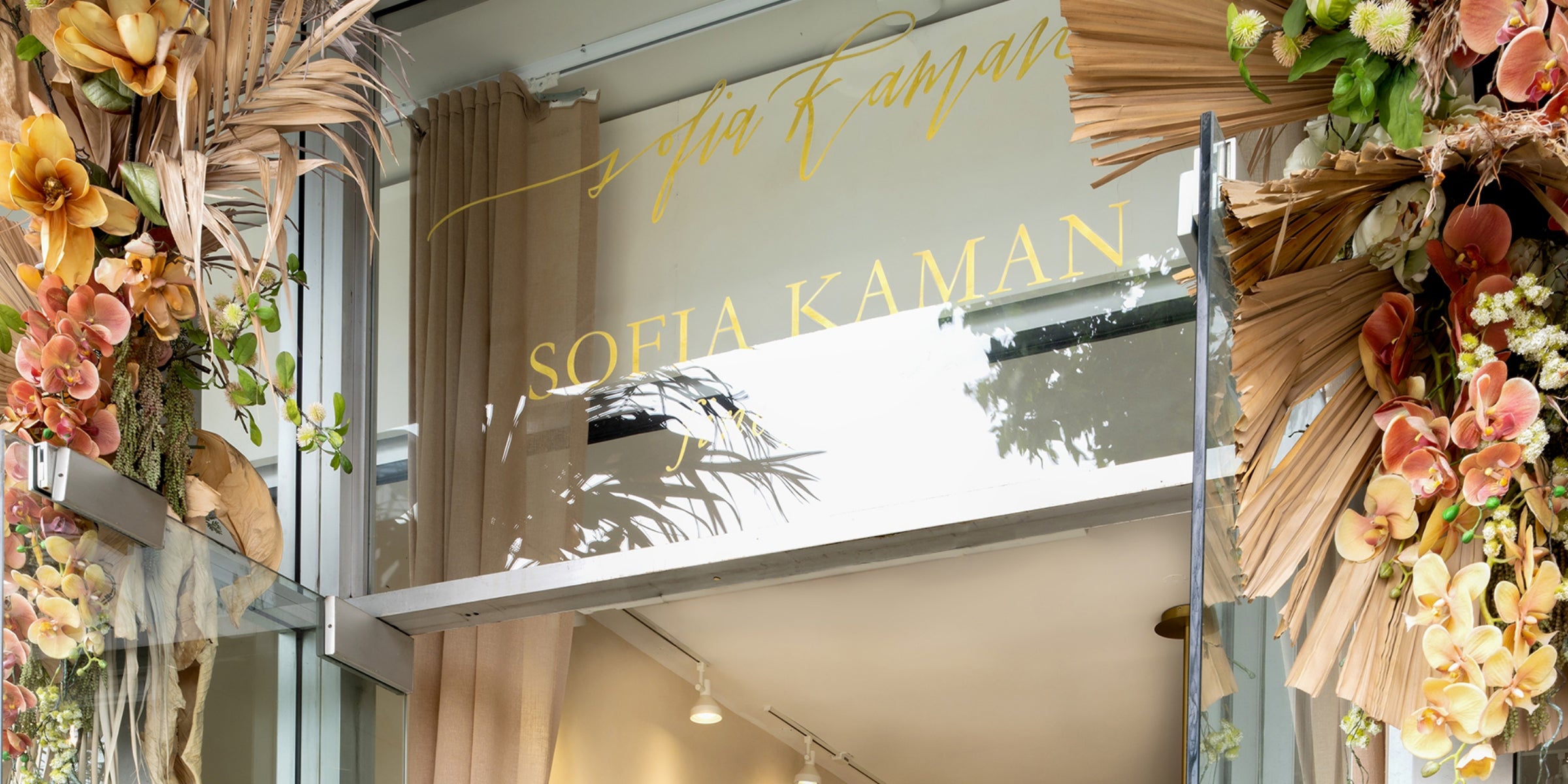 Storefront entrance with hanging peach and yellow flowers and a gold sign reading SOFIA KAMAN