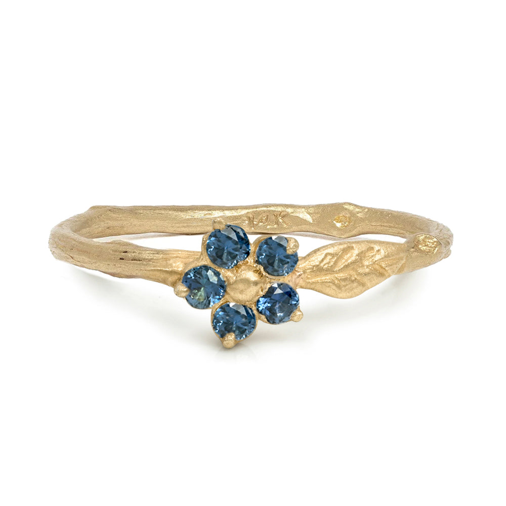 A gold ring featuring a textured band with leaf-like details and a central flower motif set with five round blue gemstones, shown on a plain white background.