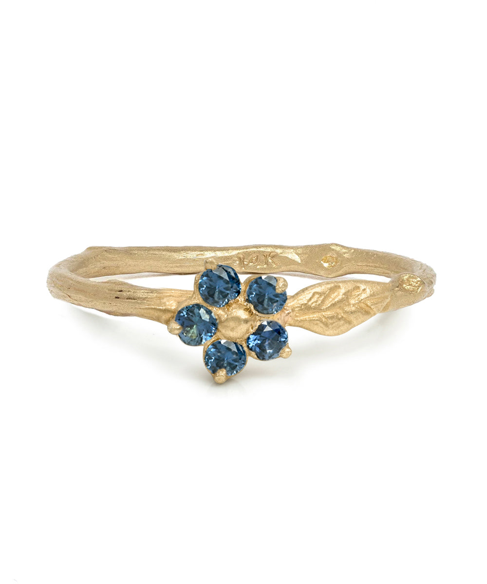 A gold ring featuring a textured band with leaf-like details and a central flower motif set with five round blue gemstones, shown on a plain white background.
