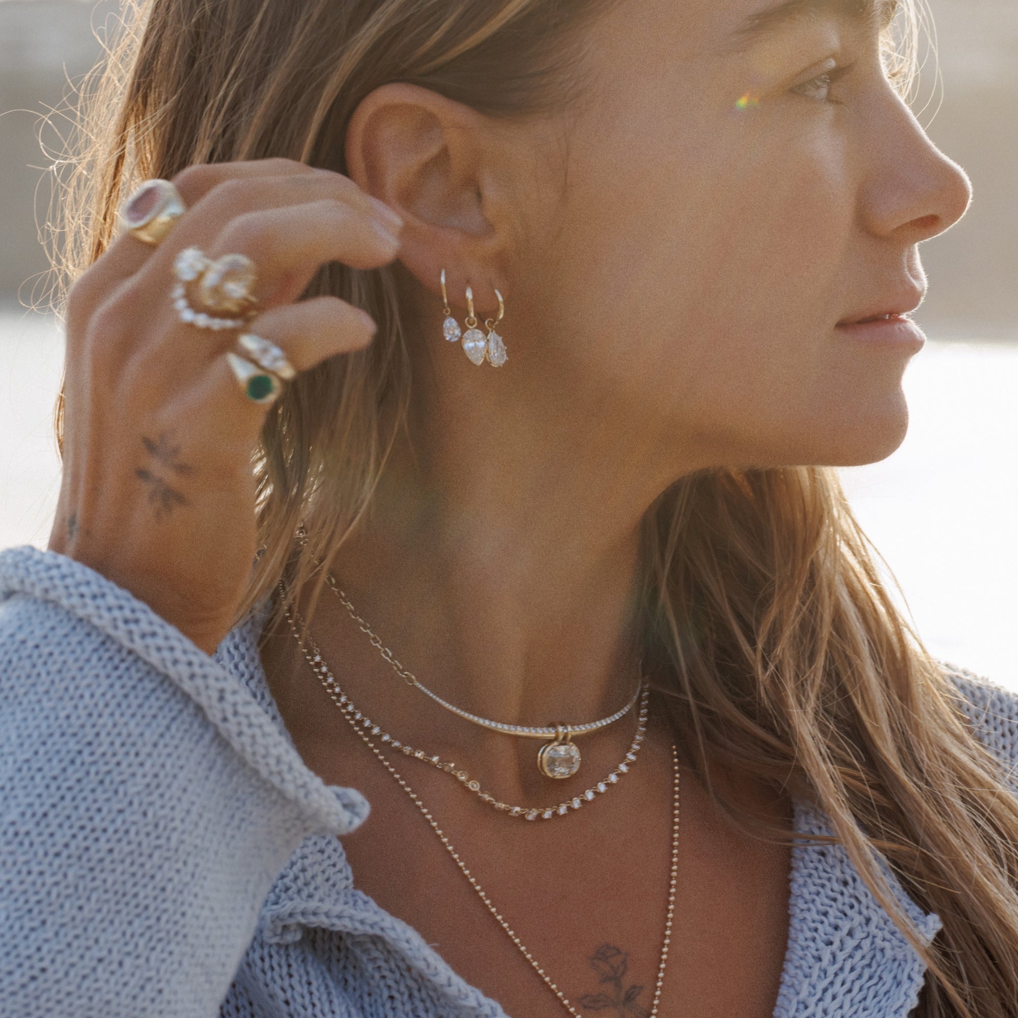 A close-up lifestyle shot of a woman wearing delicate gold-toned layered necklaces, a silver chain choker with a small heart pendant, and matching small pearl drop earrings. She is also wearing multiple unique engagement rings designed by Sofia Kaman on her fingers, including one with a green stone, against a softly blurred outdoor background.