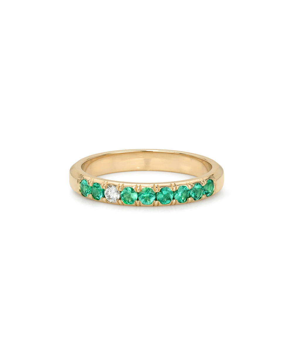 A yellow gold eternity ring featuring a row of round-cut green emeralds with one central clear diamond, set against a plain white background.