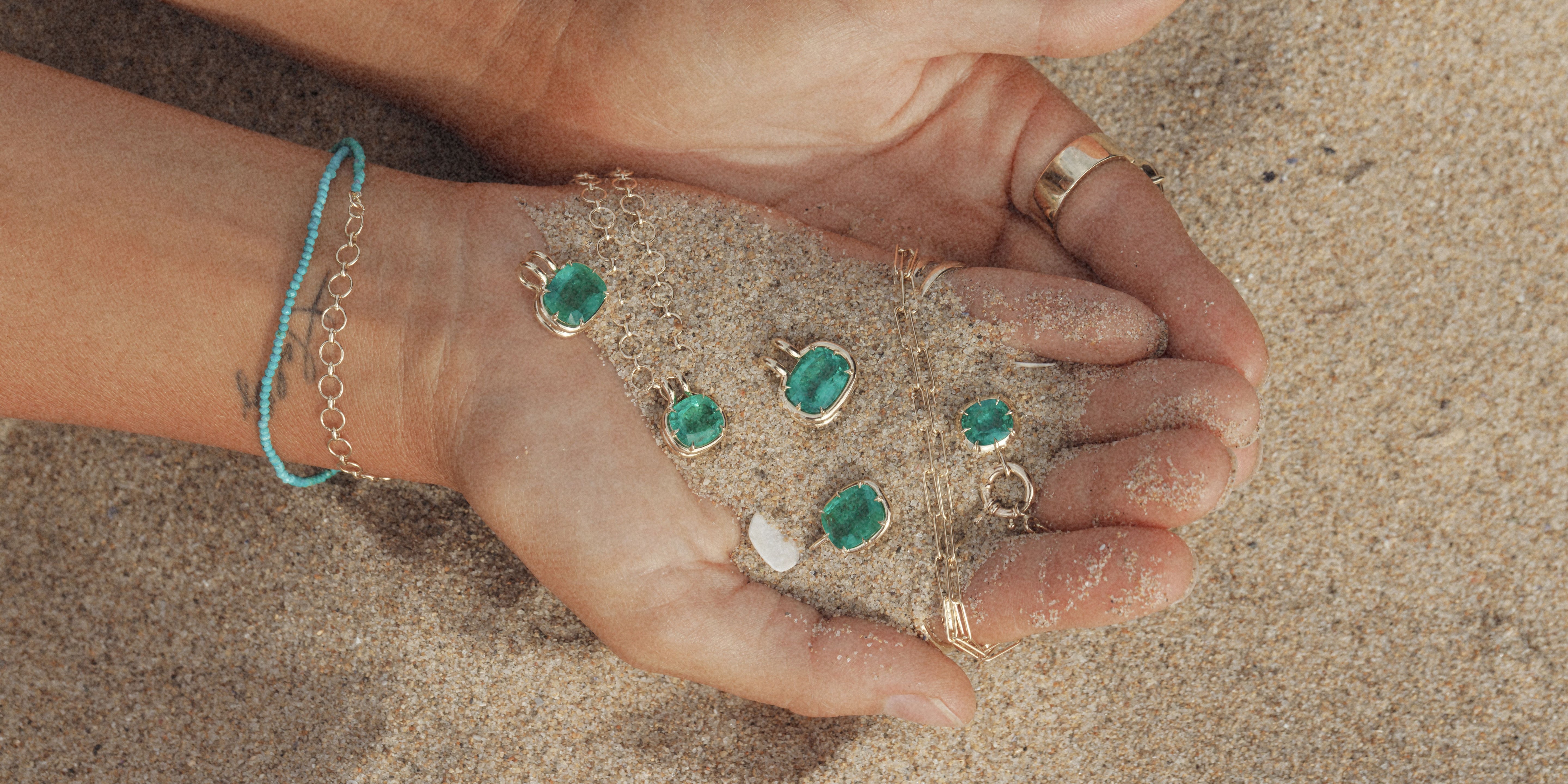 A close-up of two hands holding sand on a beach, with several gold-tone jewelry pieces featuring green oval stones displayed on the sand. A turquoise chain bracelet is also visible on the wrist.