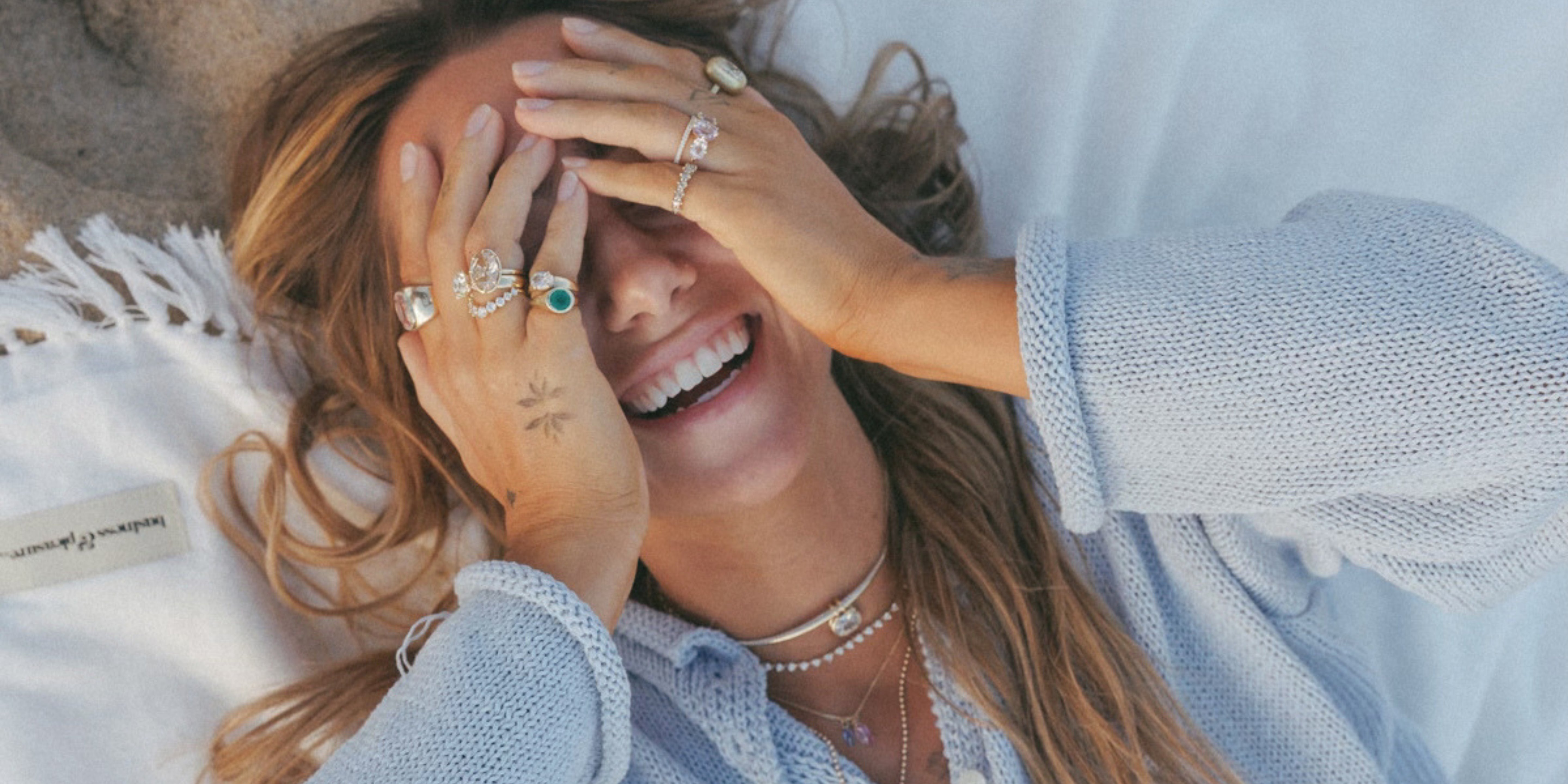 A woman lying on a white blanket outdoors, smiling and covering one eye with both hands, which are adorned with multiple silver rings.