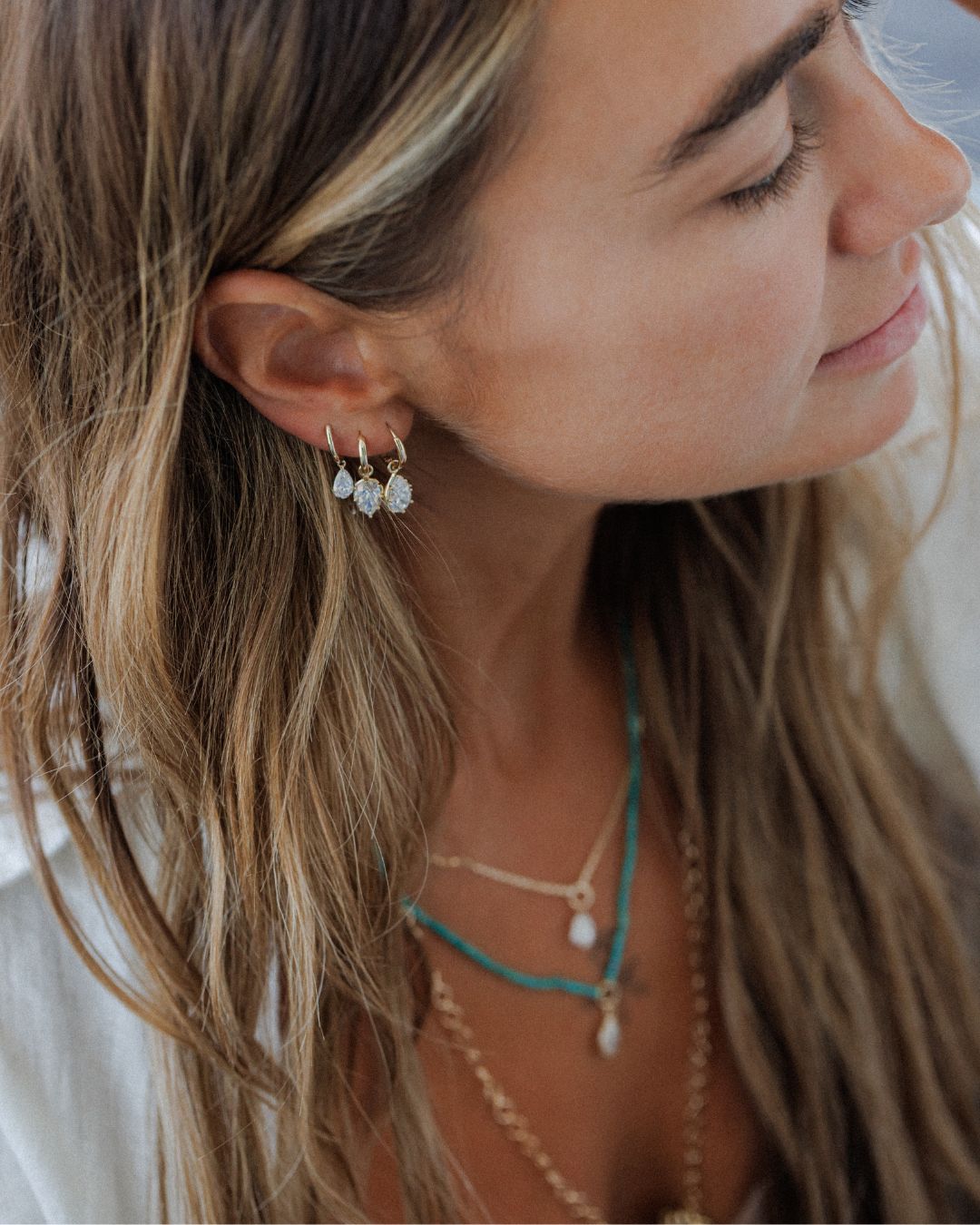A close-up side profile of a woman with long light brown hair wearing small gold hoop earrings with white teardrop-shaped crystals, layered with turquoise and gold necklaces.