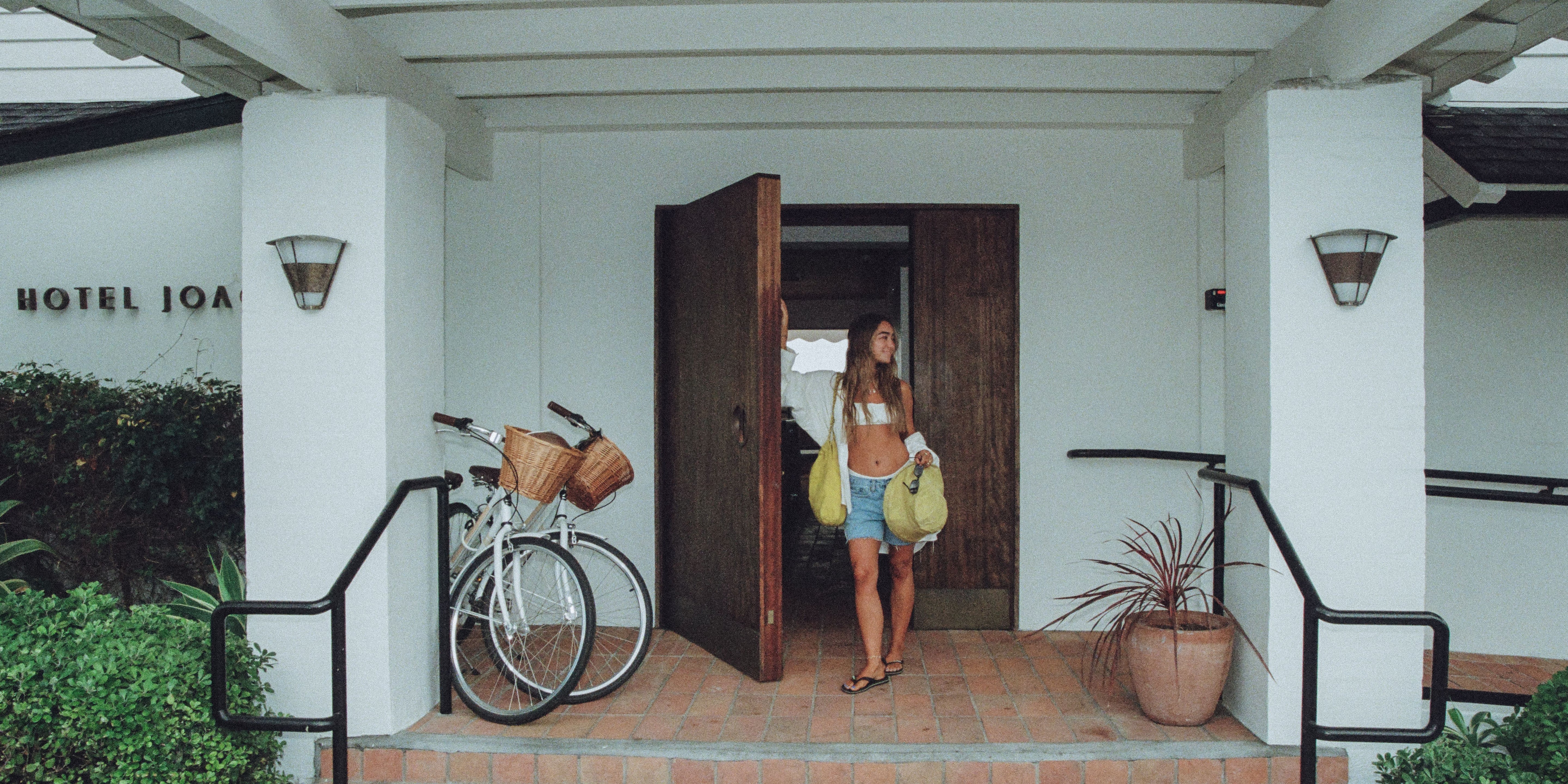 A woman stands in the open doorway of a white hotel, carrying two yellow bags, with a white bicycle parked on the left and a potted plant on the right.