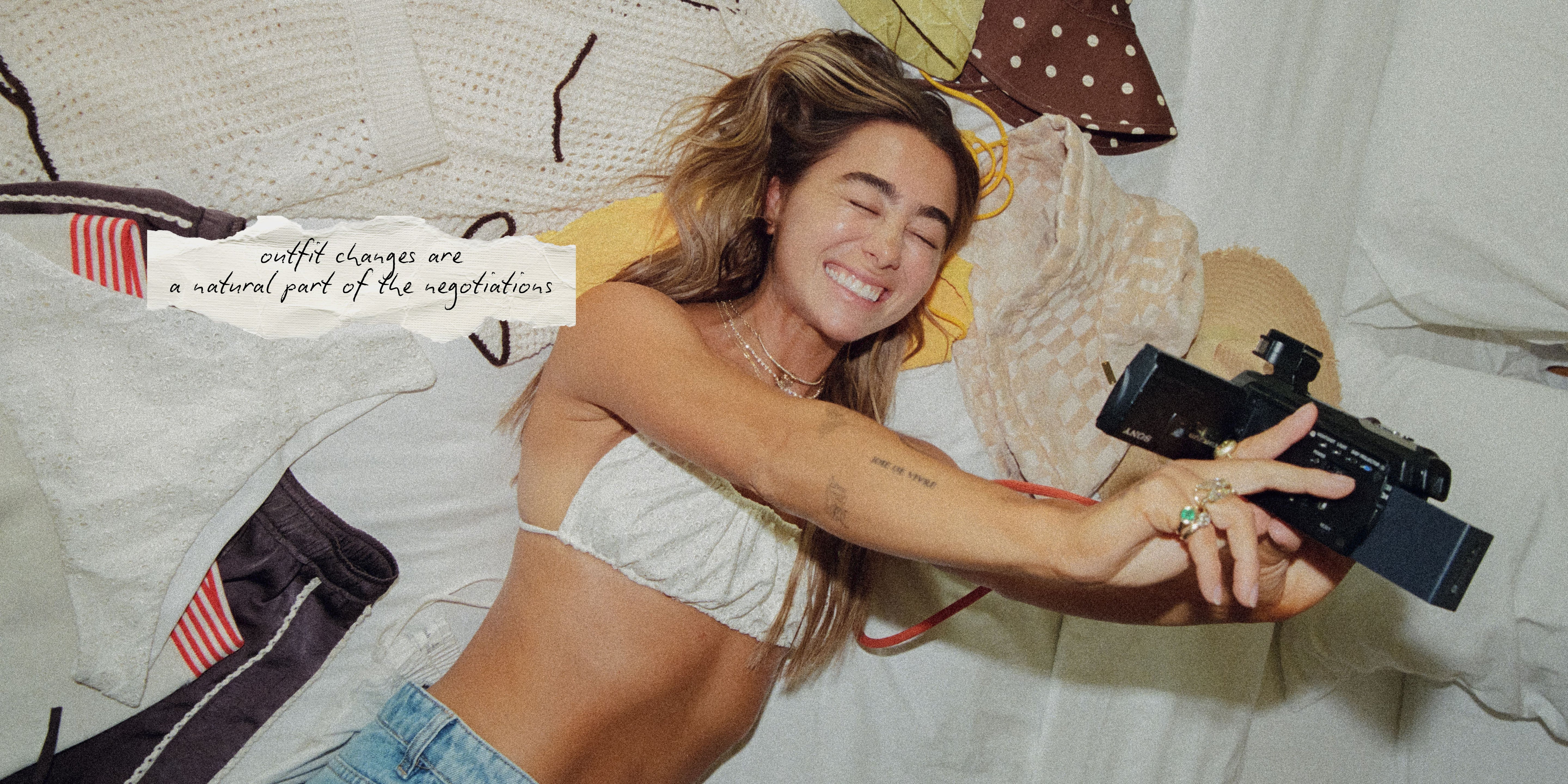 A woman lying on a bed wearing a white ribbed tube top and blue jeans, holding a black camera. A small paper tag on the left reads, 'outfit changes are a natural part of the negotiation.