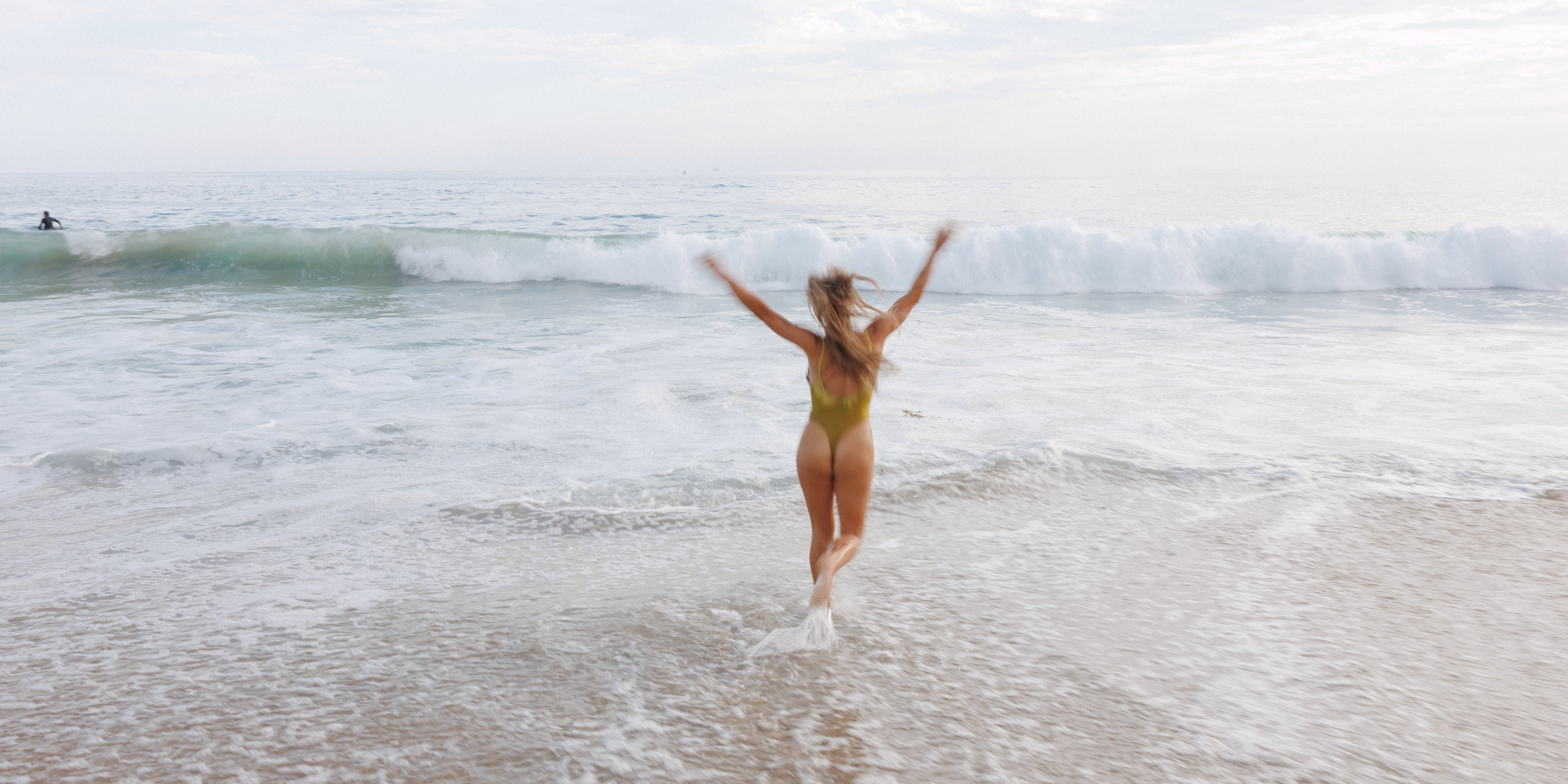 A woman in a yellow-green swimsuit runs into the ocean waves with her arms raised, standing on a sandy beach under an overcast sky.