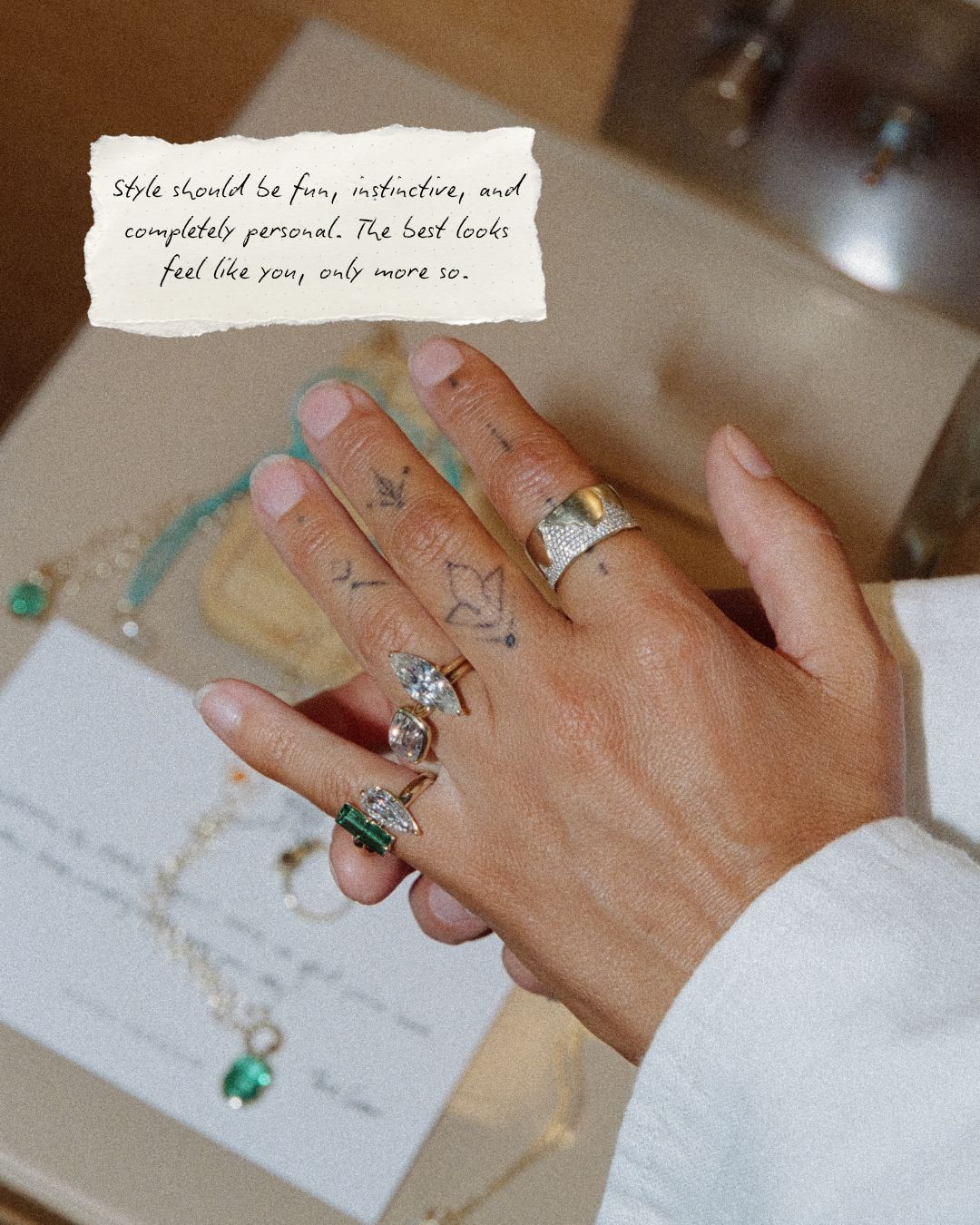 A close-up of a person's hand wearing multiple rings, including gold rings with diamonds and a green stone ring, with a beige card and jewelry pieces in the background. A torn paper note contains handwritten text.