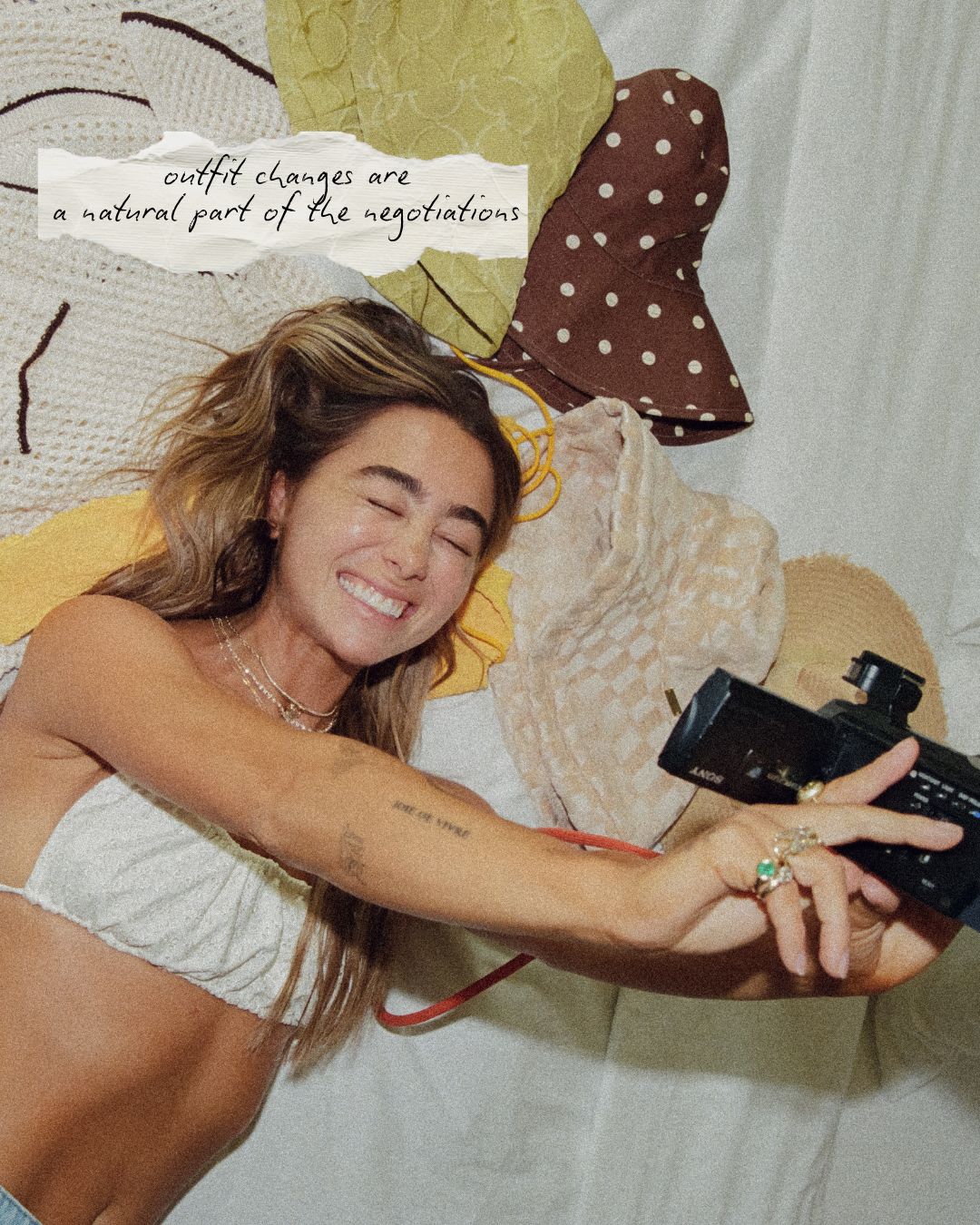 A woman lying on a bed wearing a white bikini top and holding a black camera, with a torn-paper style text overlay that reads 'outfit changes are a natural part of the negotiations'. She is surrounded by a yellow and white patterned blanket and a brown polka-dot sun hat.