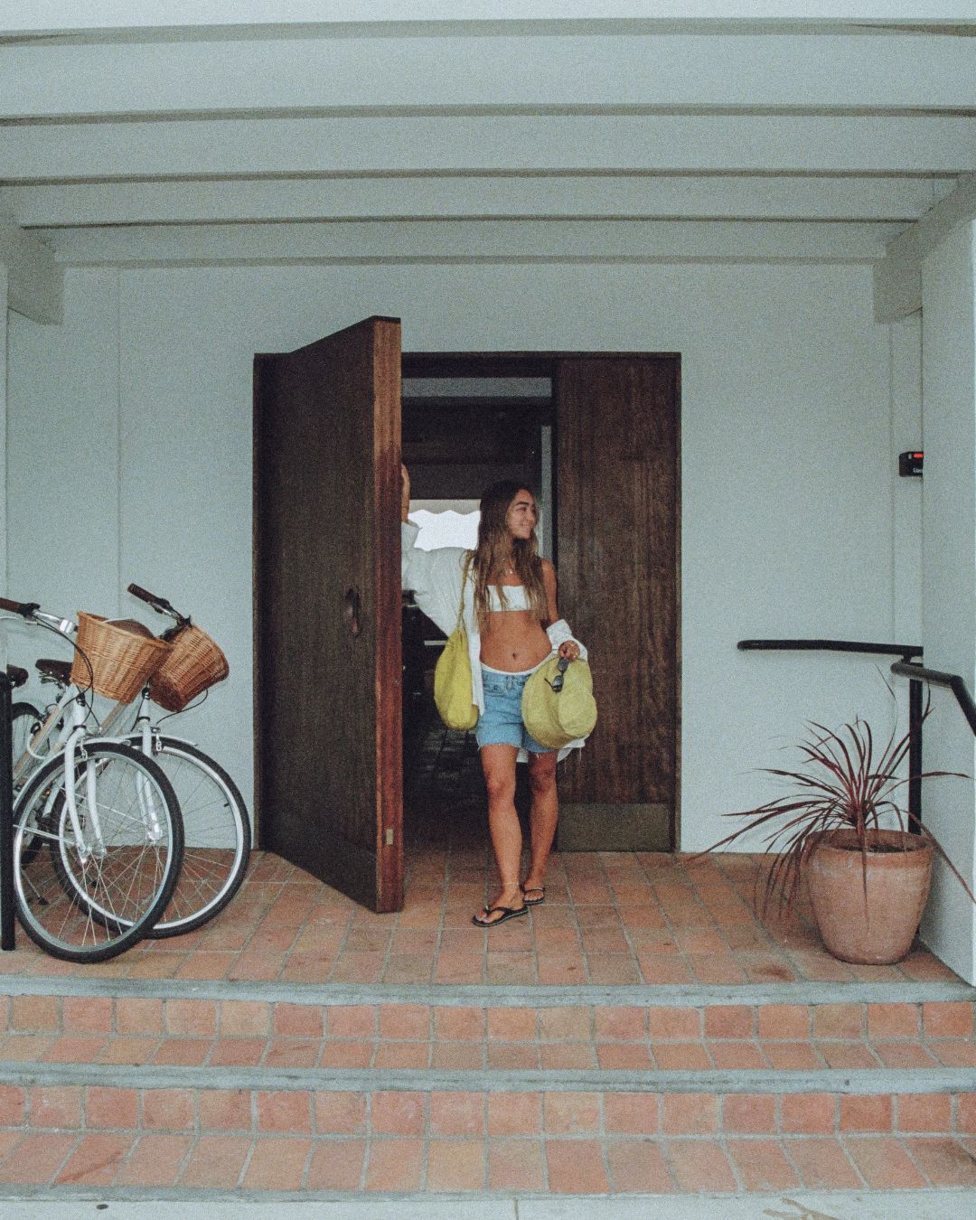 A woman stands in a doorway holding two yellow bags, with a white bicycle on the left and a potted plant on the right, set against a white stucco wall and terracotta steps.