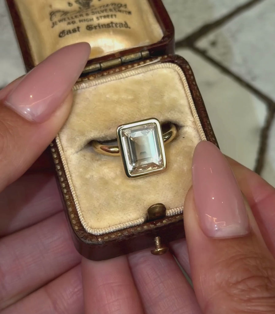 A yellow gold ring featuring a large rectangular portrait cut clear champagne diamond, shown in a vintage ring box and hands modeling.