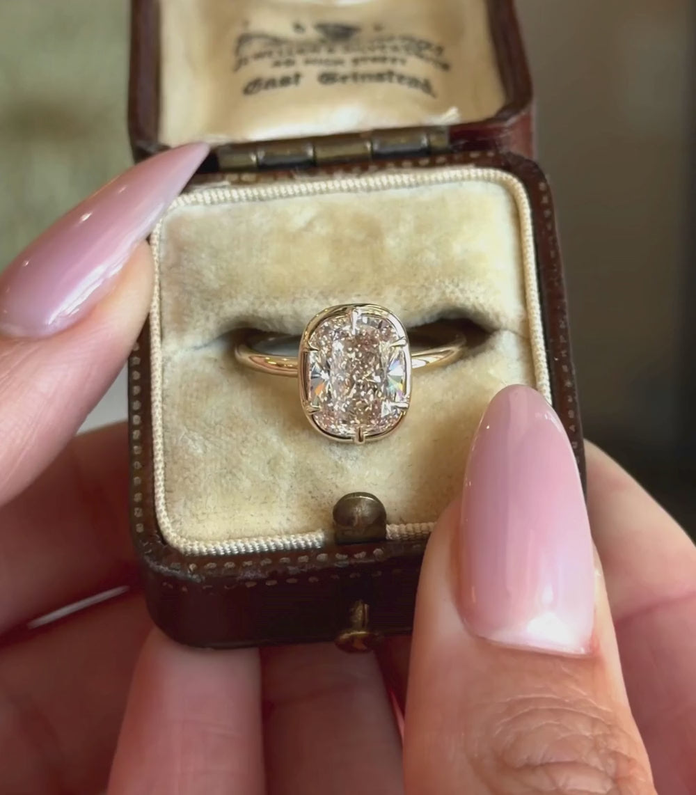 A yellow gold ring featuring a cushion shaped brilliant cut champagne diamond in a six prong setting and faux bezel, shown in a vintage ring box and hands modeling.