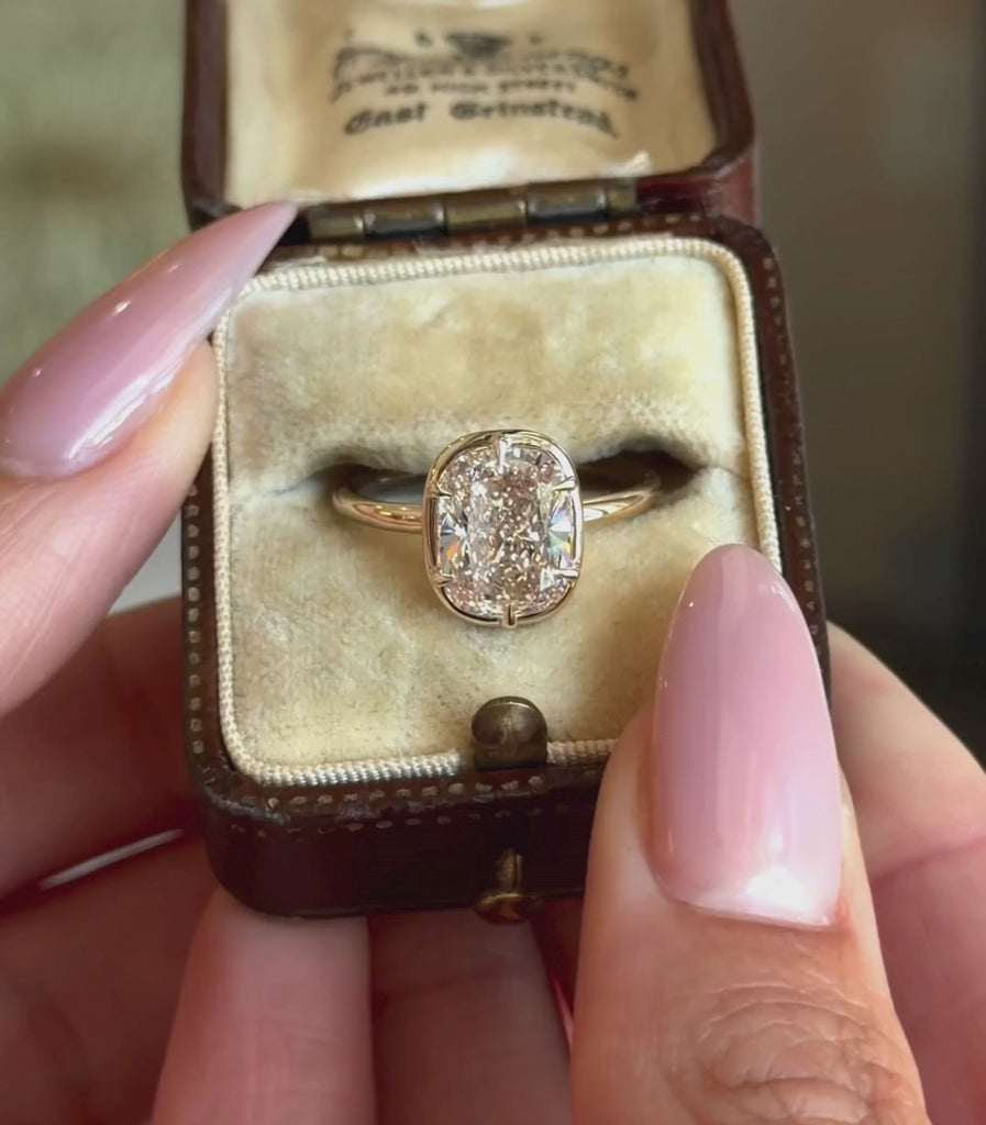 A yellow gold ring featuring a cushion shaped brilliant cut champagne diamond in a six prong setting and faux bezel, shown in a vintage ring box and hands modeling.