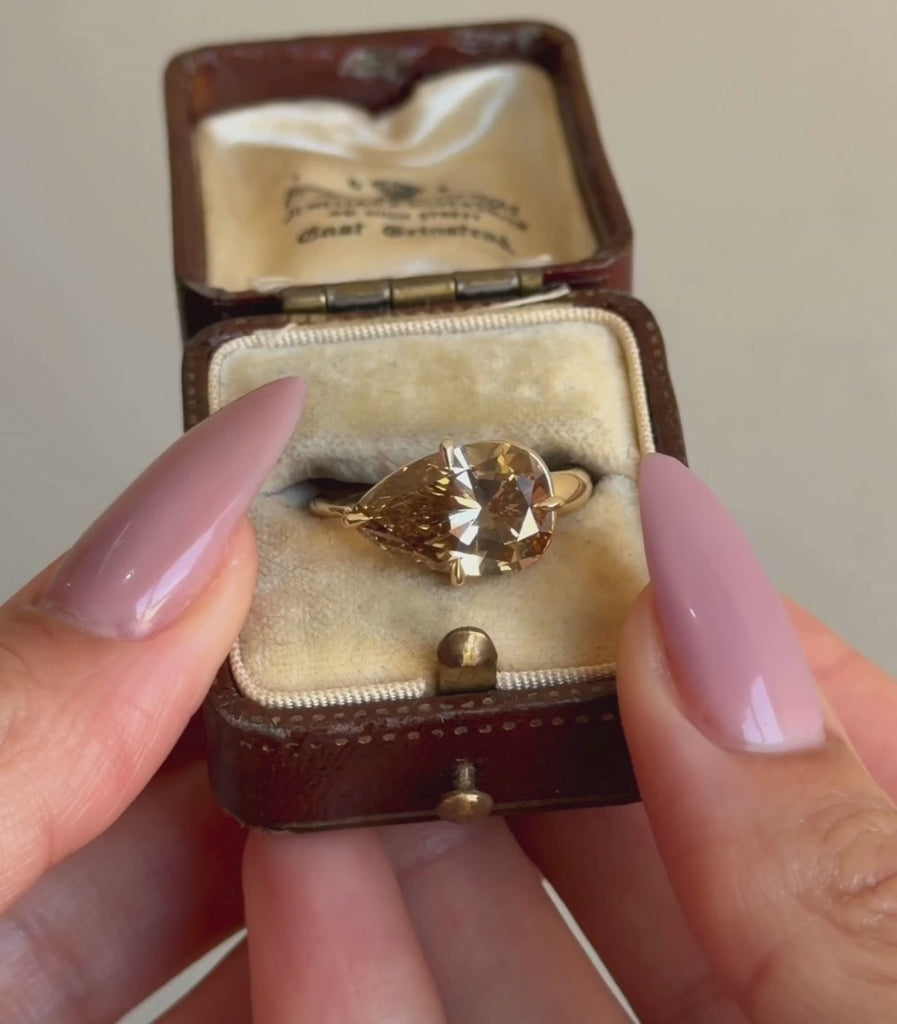 14K yellow gold ring with a 4.52ct pear shaped brilliant cut champagne diamond set east west in a engagement ring setting shown in a box with hands.