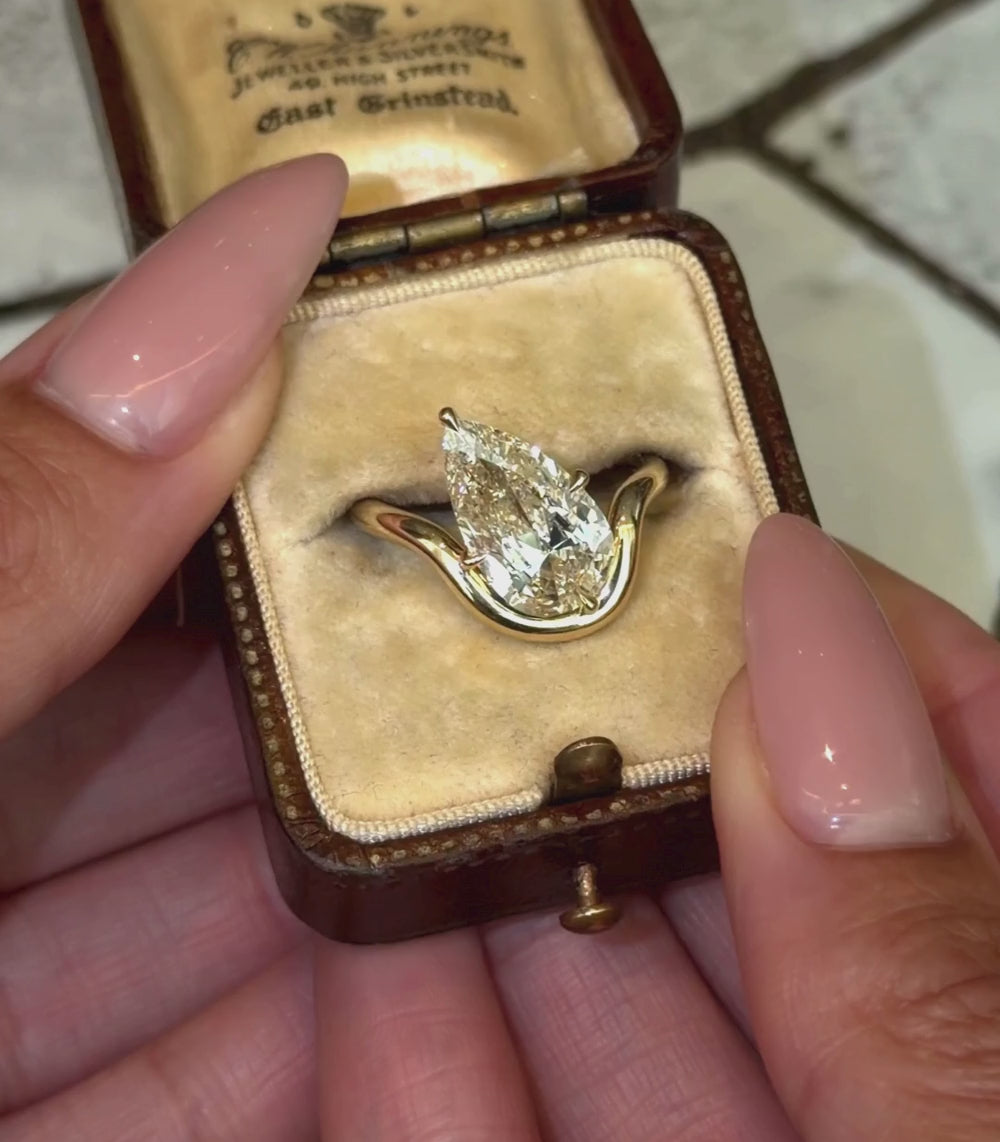 A pear-cut diamond yellow gold ring in a yellow gold setting with a wavy, sculptural band, shown in a vintage ring box and with hands modeling.