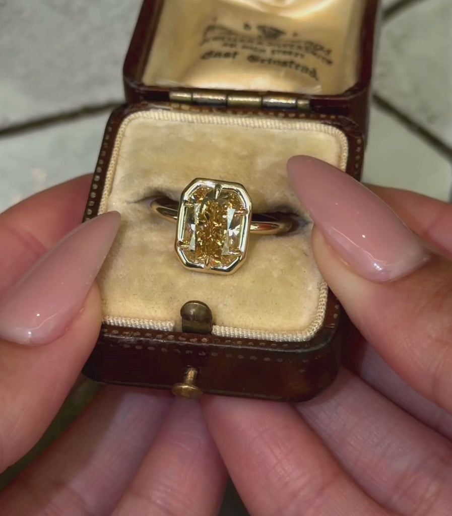 A yellow gold ring featuring a radiant shaped brilliant cut champagne diamond in a six prong setting and faux bezel, shown in a vintage ring box and hands modeling.