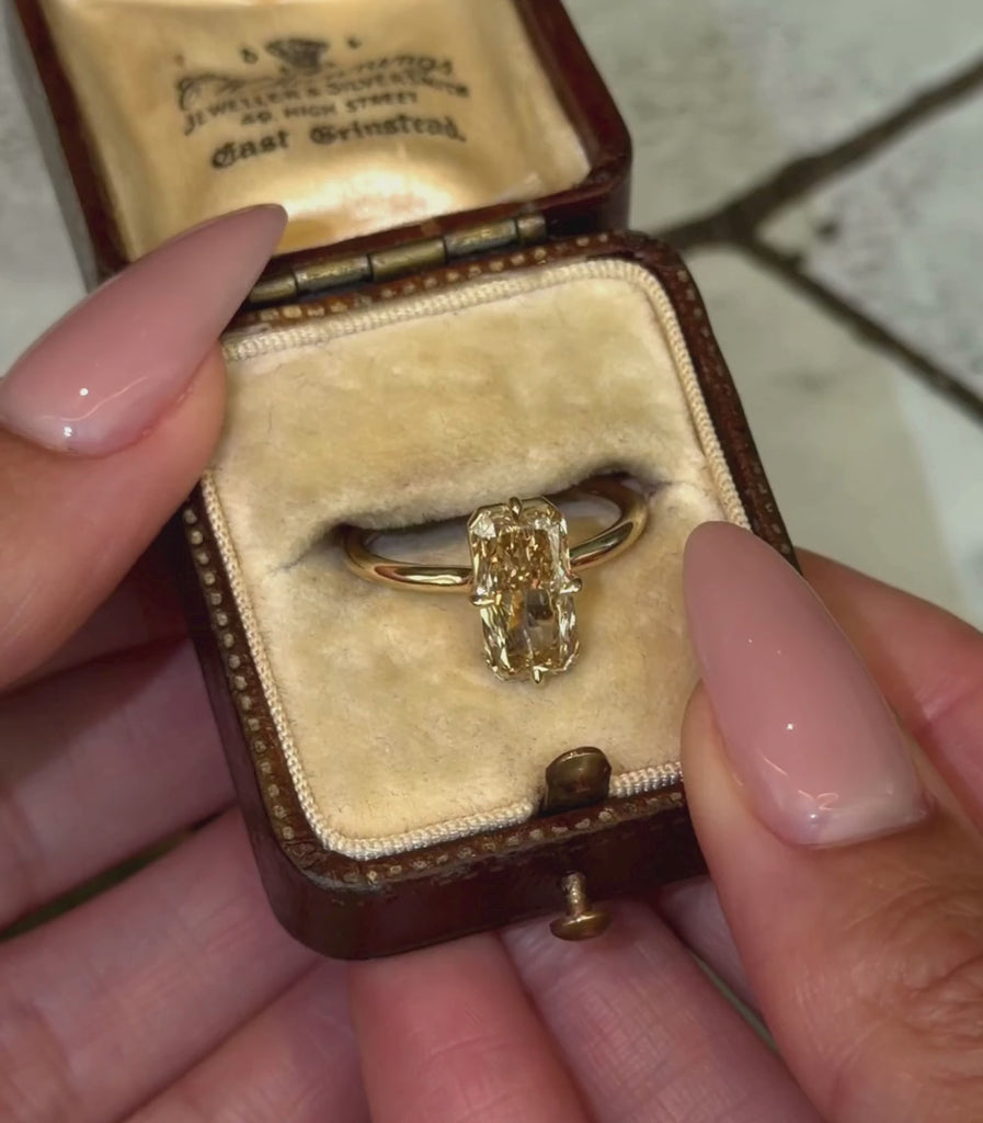 A yellow gold ring featuring a large rectangular radiant brilliant cut champagne diamond in a 4 prong setting, shown in a vintage ring box and hands modeling.