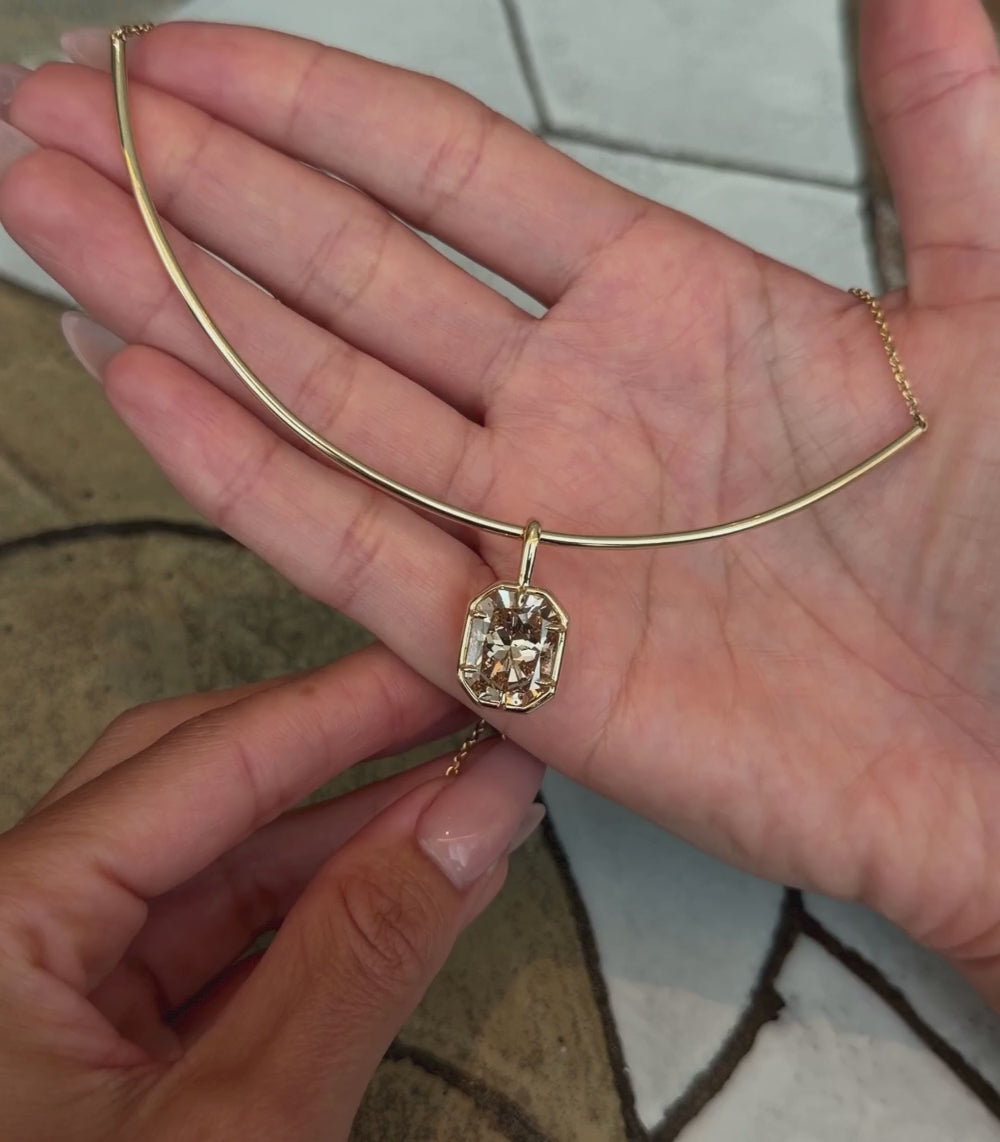 A yellow gold half collar necklace featuring a large radiant shaped brilliant cut champagne diamond pendant in a six prong setting and faux bezel, shown with hands modeling.