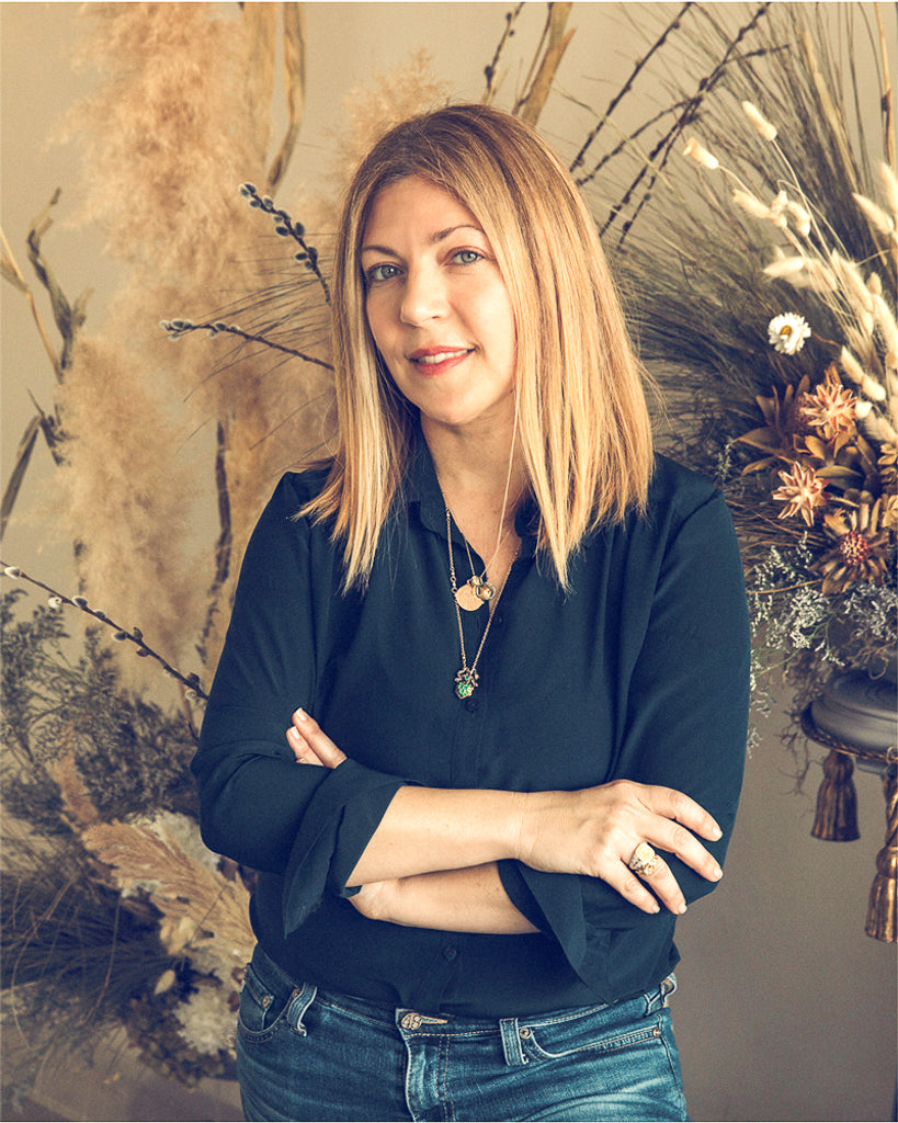 Woman in dark blouse with crossed hands displays an understated unique engagement ring beside winter florals.