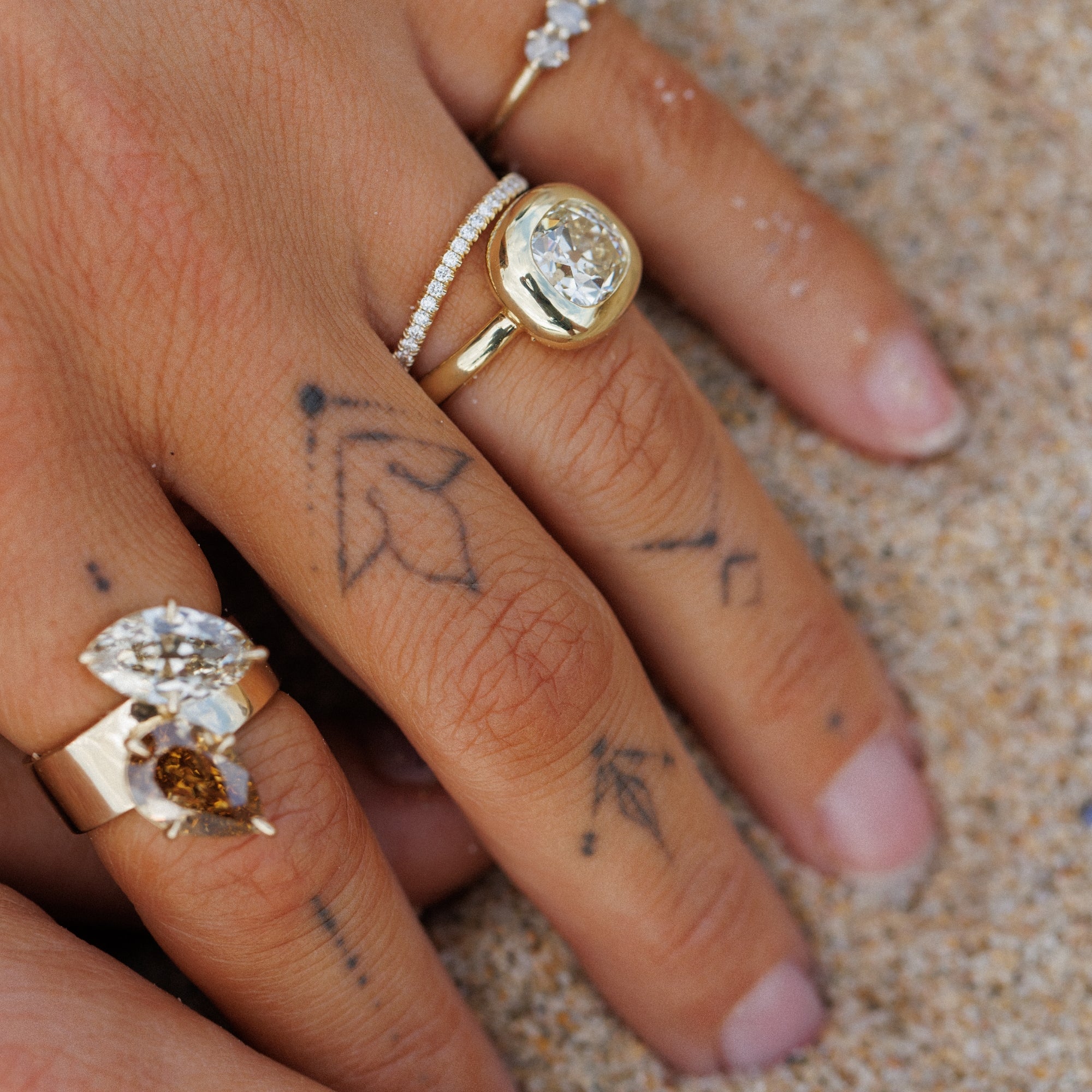 A close-up of a hand resting on sand, wearing multiple rings. The most prominent ring is a yellow gold band with a bezel-set round clear gemstone that makes the perfect unique engagement ring.