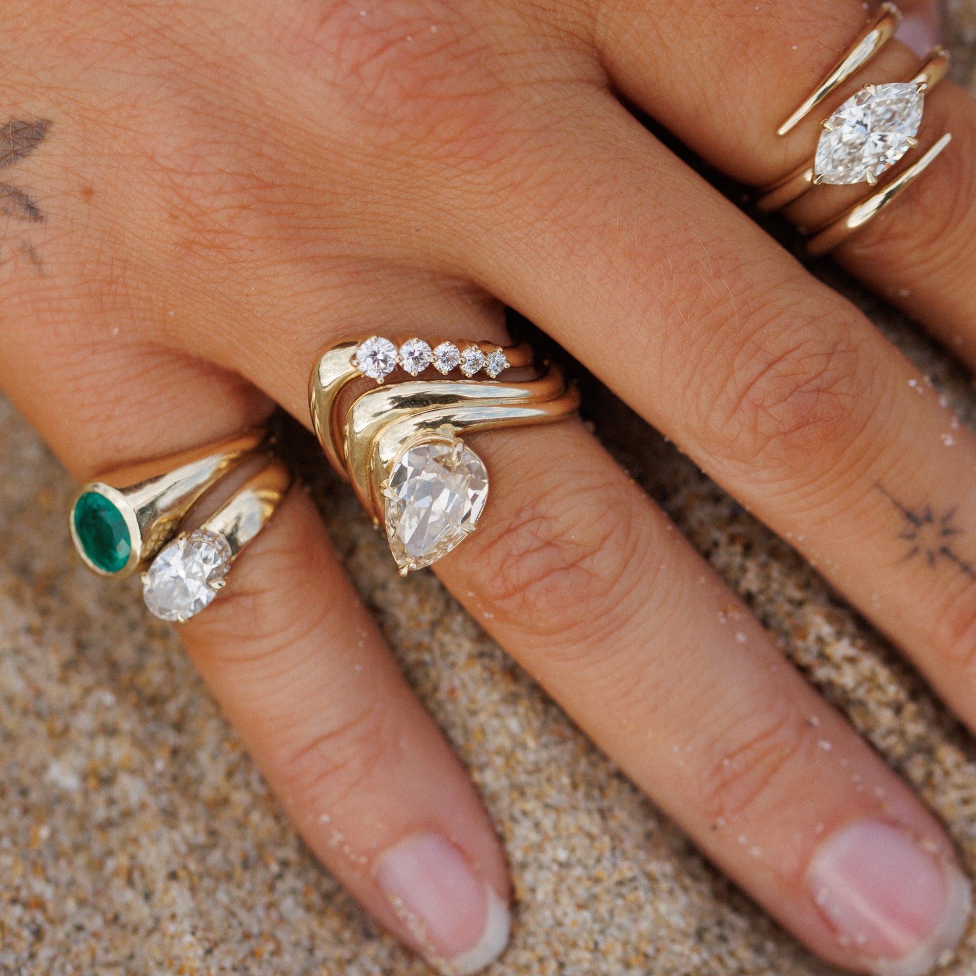 A close-up of a hand wearing multiple gold rings on a sandy surface. The rings include a marquise-cut clear gemstone, a pear-cut clear gemstone with small clear stones, and a green oval gemstone, all which make perfect unique engagement rings.