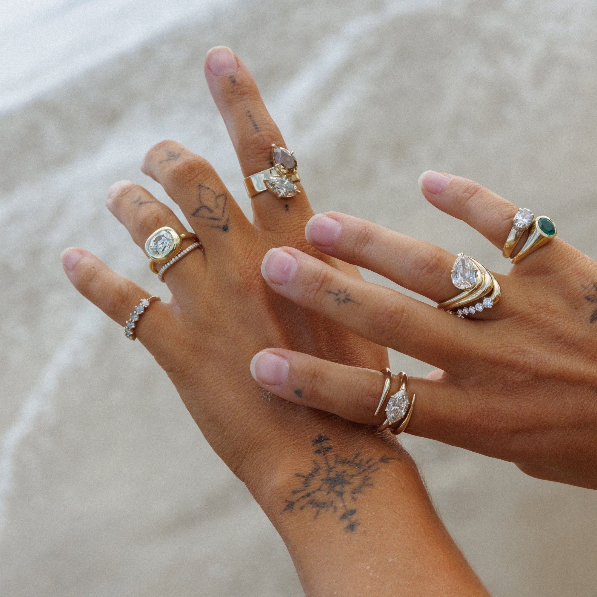 A close-up of two hands wearing multiple unique engagement rings designed by Sofia Kaman with clear stones and one green stone ring, photographed outdoors against a soft sandy background.