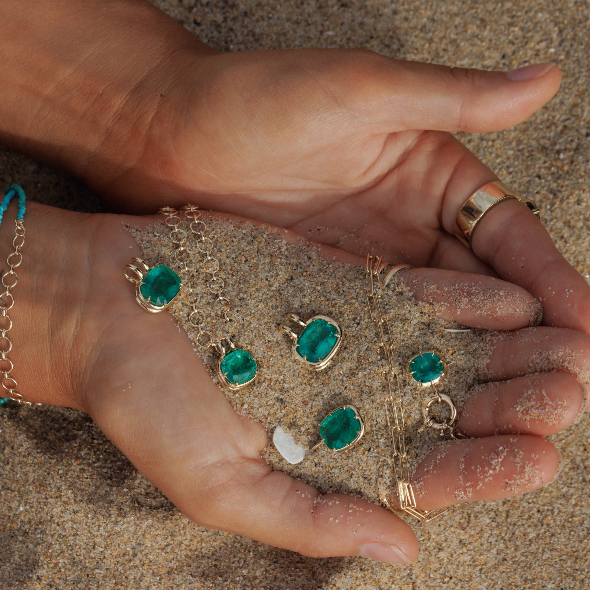 Gold-toned jewelry pieces with green gemstones are displayed on sand held in two hands, with a matching green and gold bracelet visible on the left wrist.