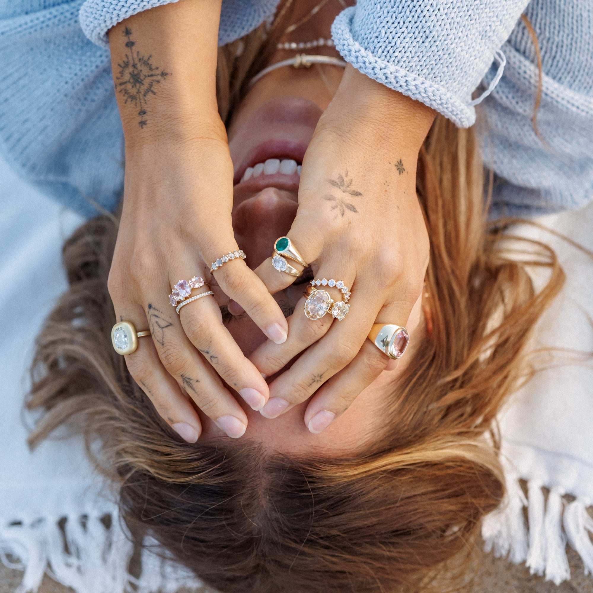 A close-up lifestyle shot of Julia Havens lying down with her hands raised to show off multiple gold rings adorned with various diamonds, emeralds and morganites, set against a soft white blanket.