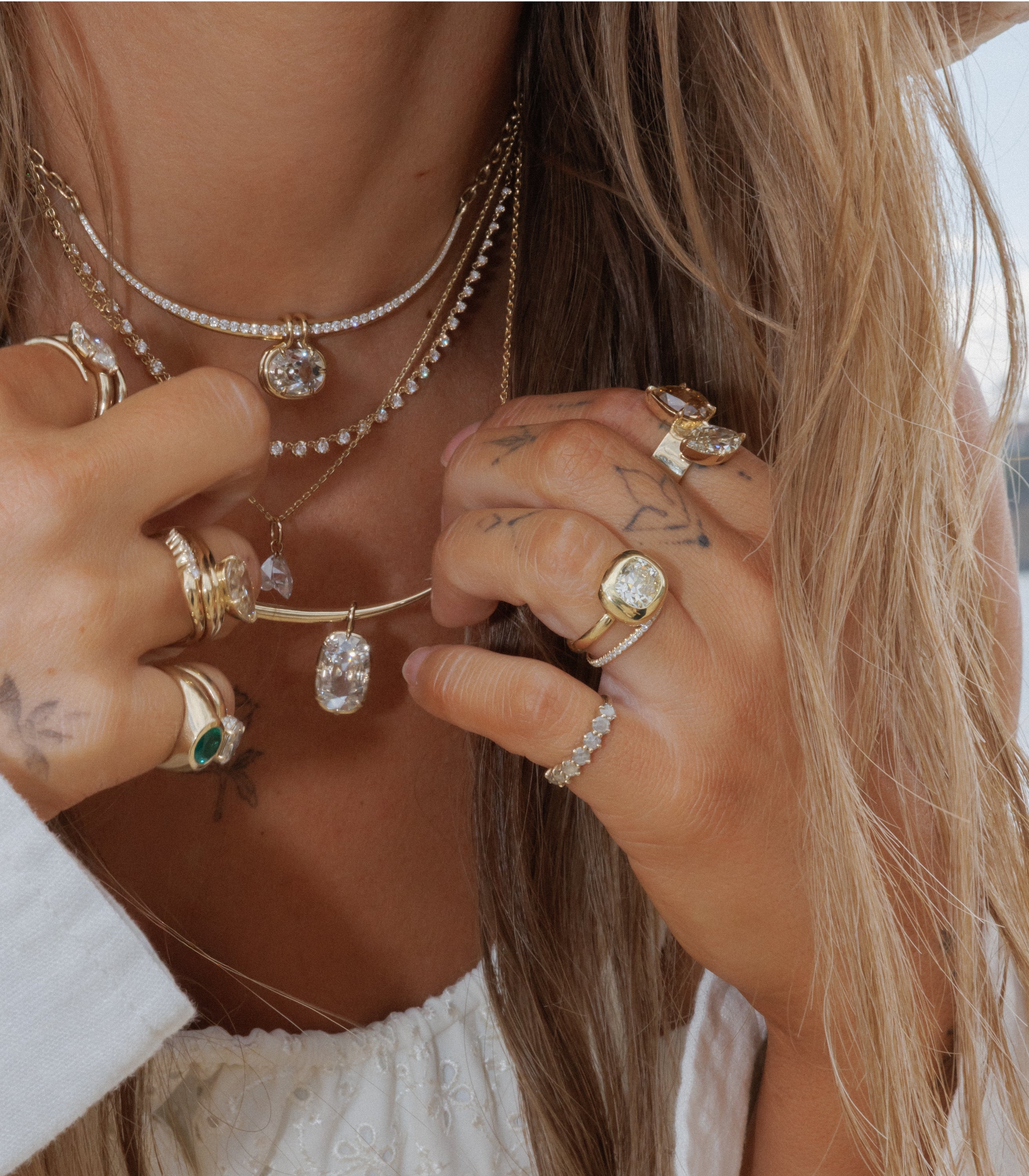 A close-up lifestyle shot of a woman wearing a layered gold necklace set with diamonds and emeralds, accessorized with multiple gold rings on her hands.