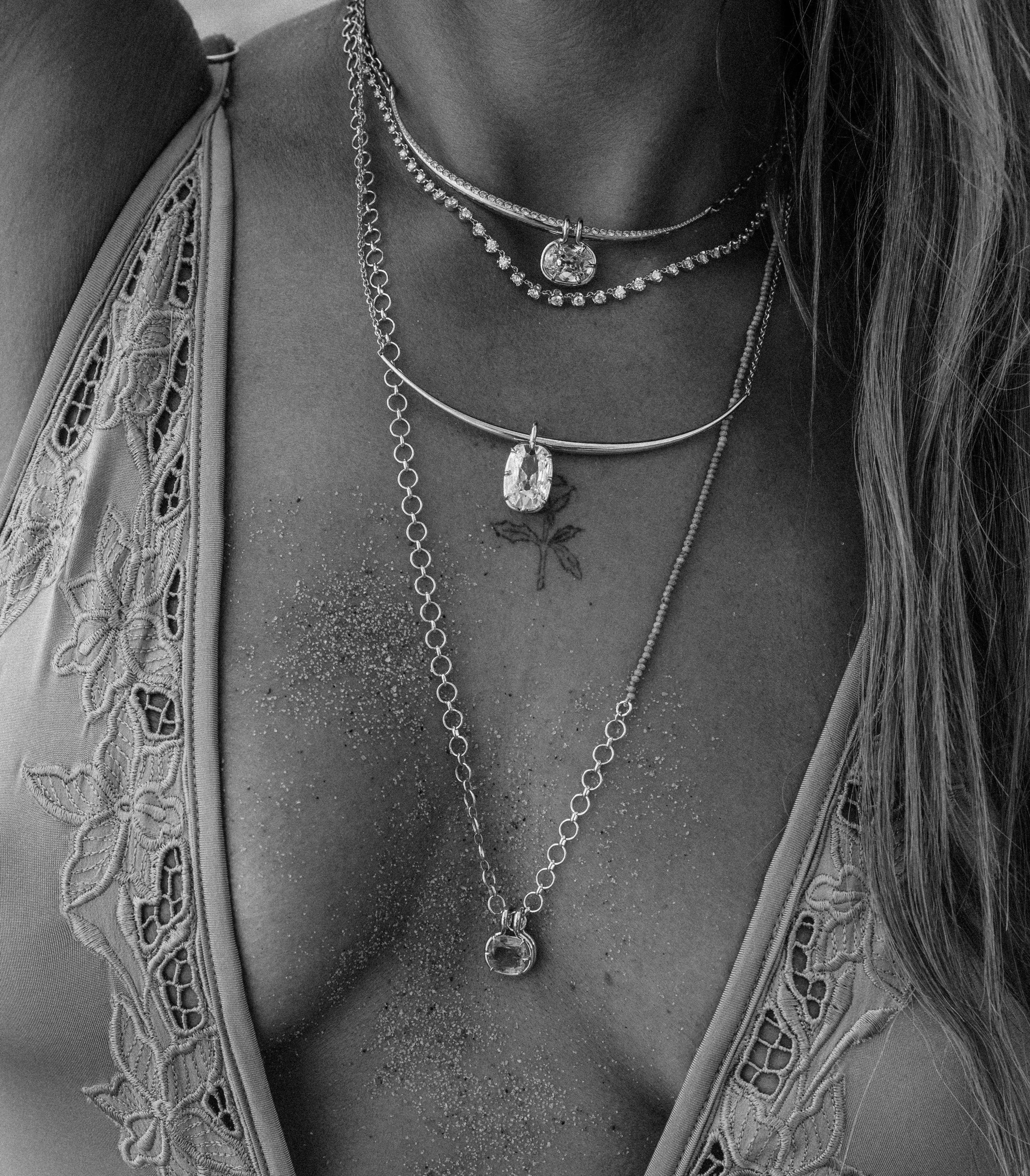A close-up black and white photograph of a woman's neck and chest wearing layered 14k yellow gold necklaces, including a diamond choker, a thin chain with a lab diamond pendant, and a longer chain with an emerald pendant.