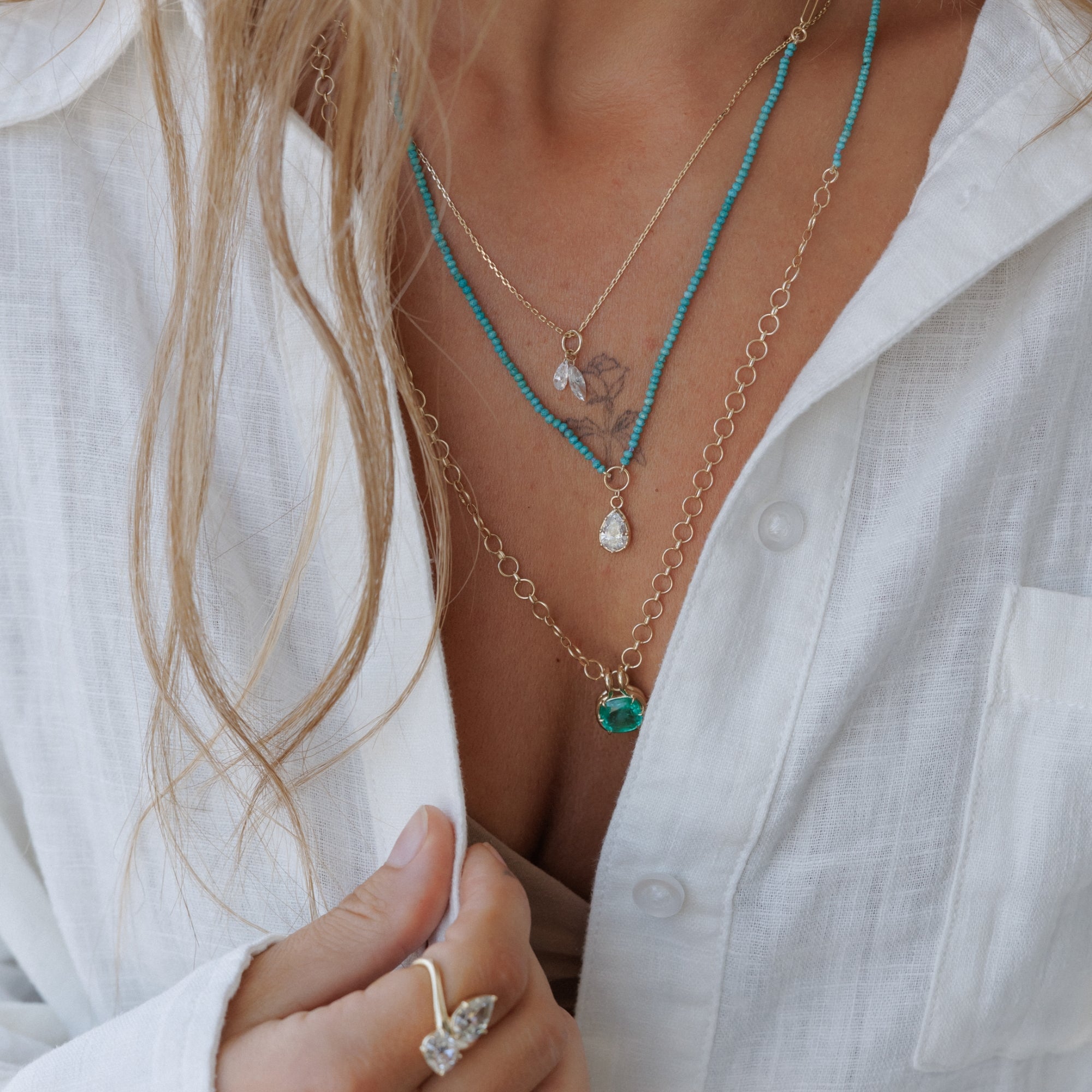A close-up of a person wearing layered necklaces over a white button-up shirt. The necklaces include a turquoise beaded chain, a gold chain with a small clear teardrop pendant, a gold chain with a small green bead pendant, and a gold chain with a small clear heart-shaped pendant.
