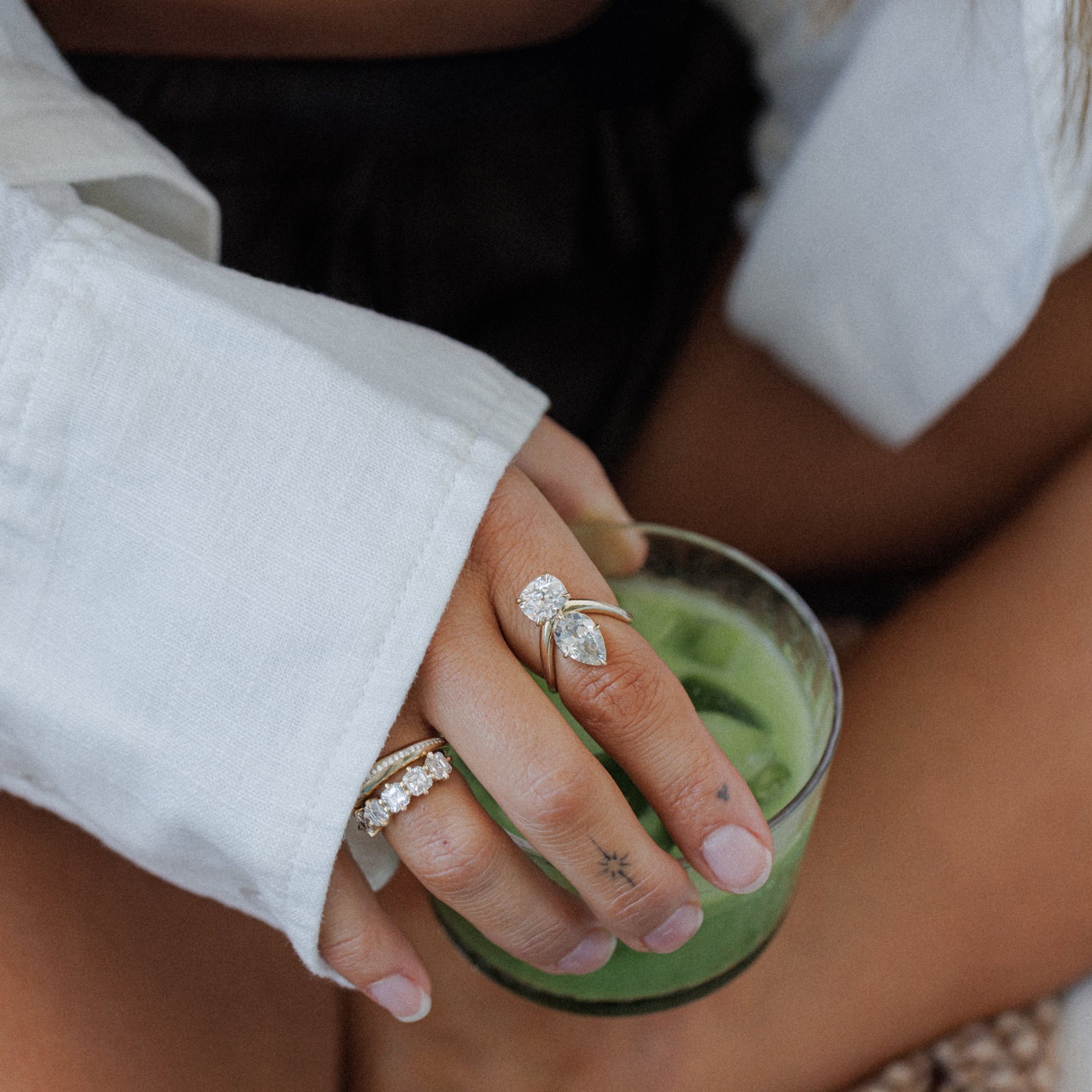 A close-up lifestyle shot of a person's hand resting on a green candle, wearing two silver rings: one with a marquise-cut clear gemstone and a smaller round clear stone, and another with a row of small clear stones, all designed by Sofia Kaman and make incredible unique engagement rings.