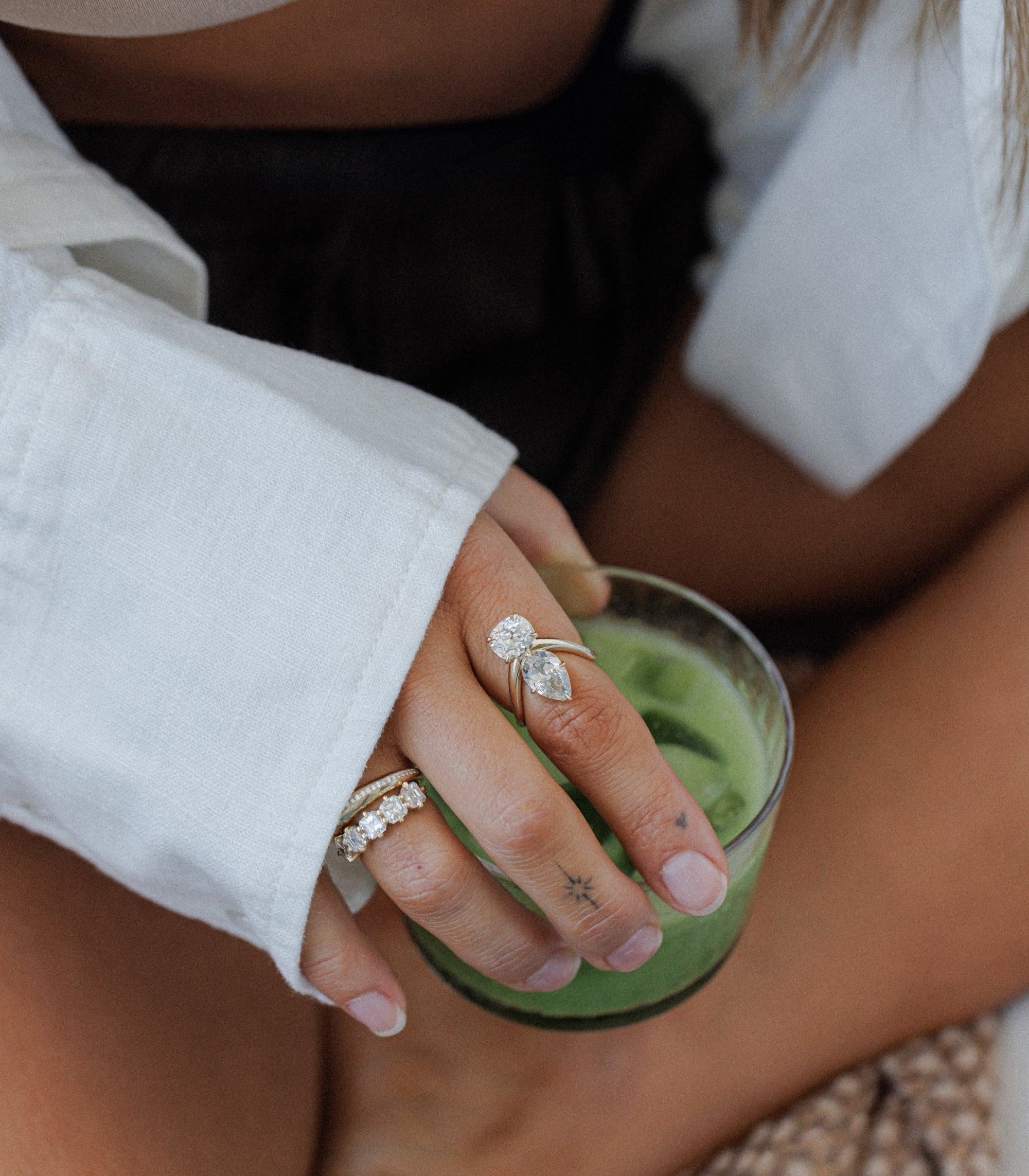 A close-up lifestyle shot of a person's hand resting on a green candle, wearing two silver rings: one with a marquise-cut clear gemstone and a smaller round clear stone, and another with a row of small clear stones, all designed by Sofia Kaman and make incredible unique engagement rings.