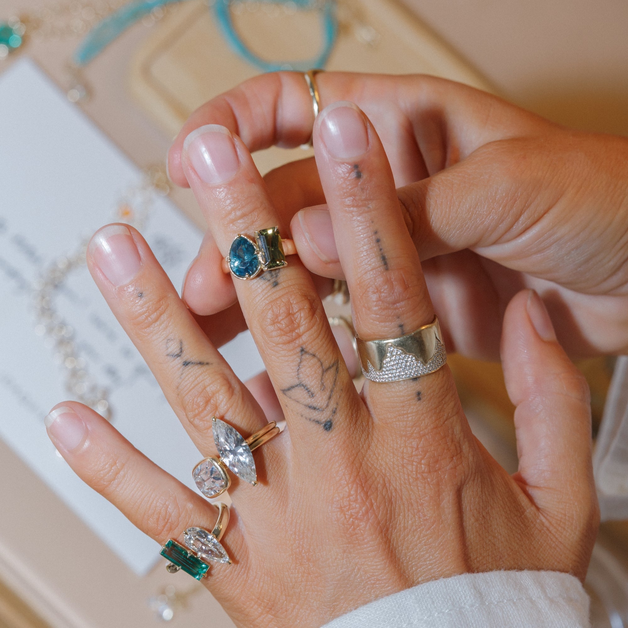 A close-up of a hand wearing multiple bohemian rings, including a textured silver band, a gold ring with a blue stone, and other silver rings with green and clear stones, all designed by Sofia Kaman  and would all make fabulous unique engagement rings.